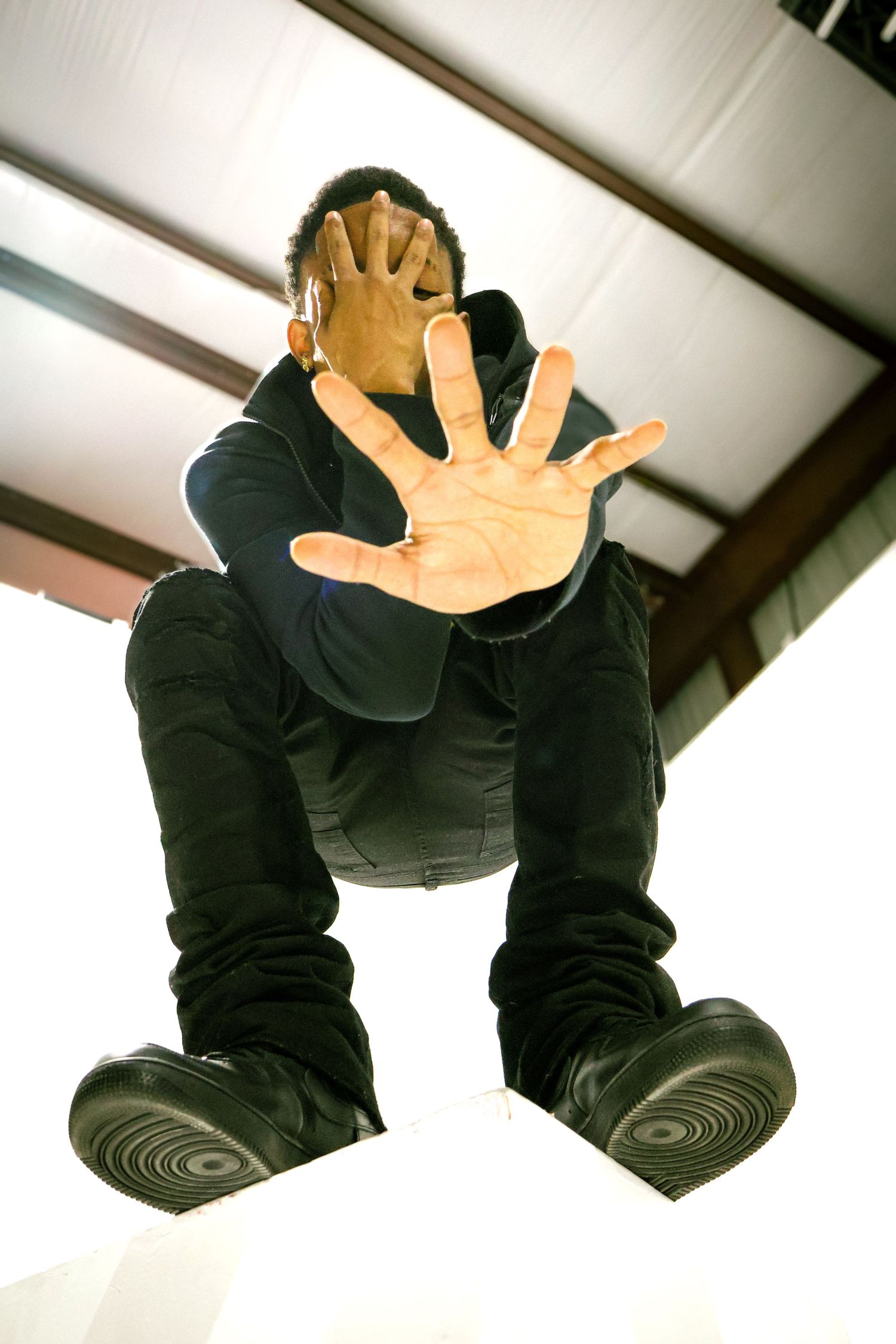 Man in black outfit crouching low, extending hand toward camera in stop gesture, shot from below ground angle.