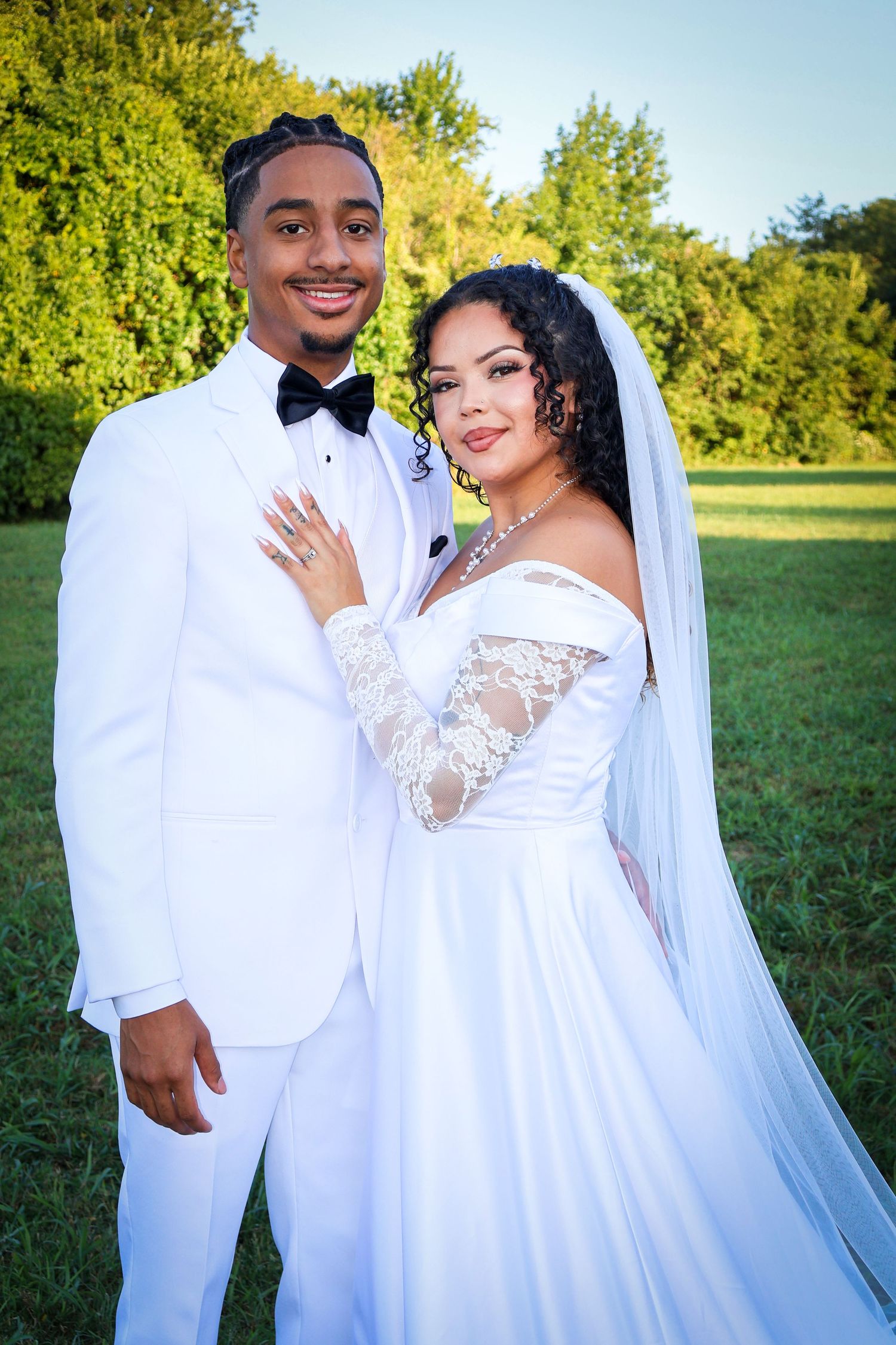 Young couple in matching white wedding attire pose outdoors on green grass with trees in background.