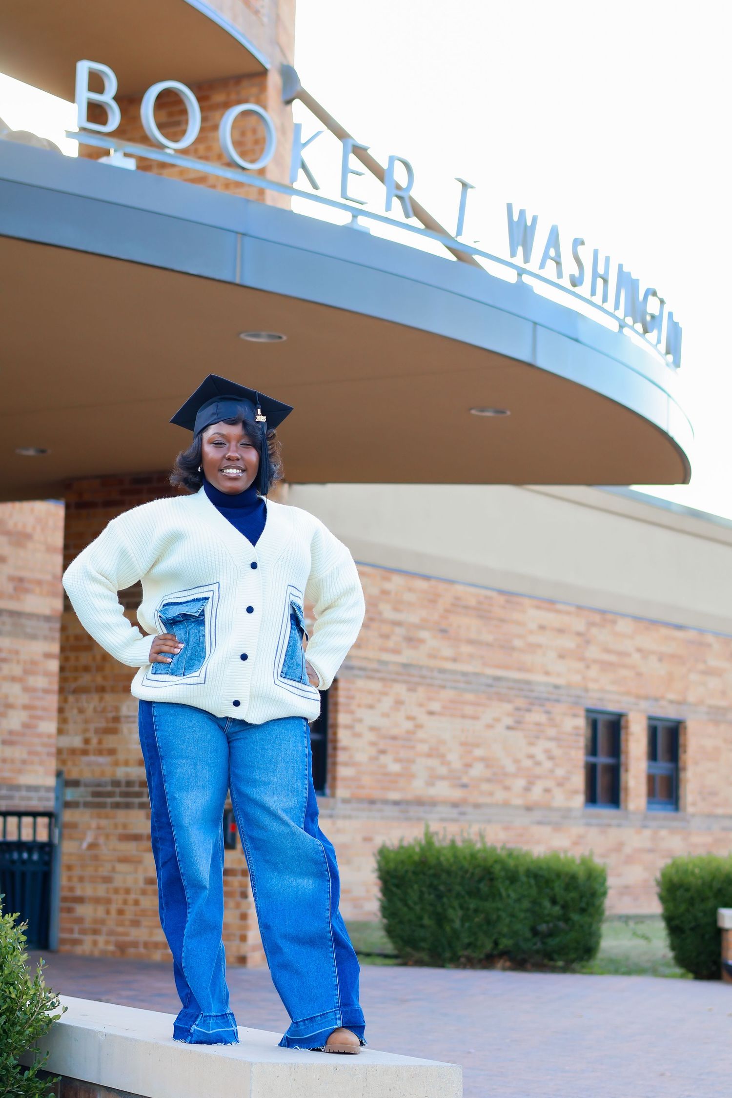 Smiling graduate in cap and cardigan posing in front of Booker T. Washington building.