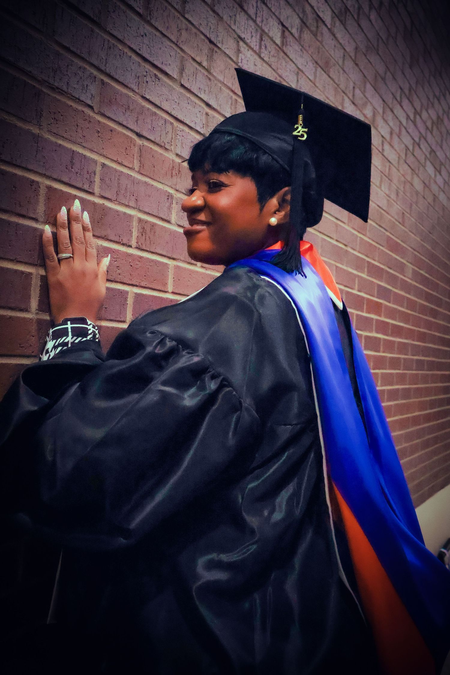 Smiling graduate in black cap and gown with blue and orange sash poses against brick wall.