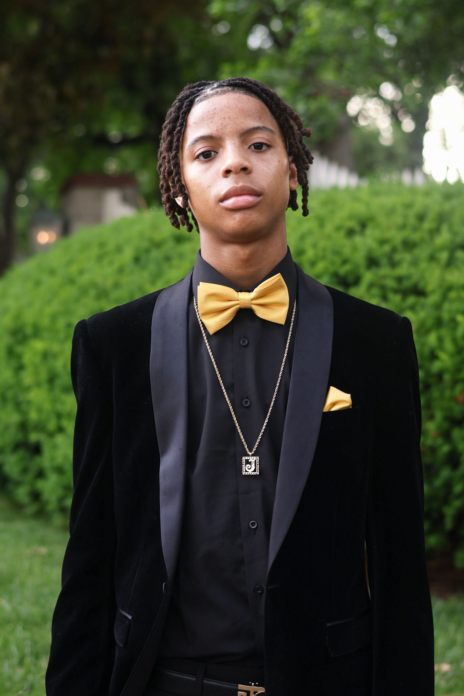 Young Black man in black velvet tuxedo with gold bow tie and chain necklace poses outdoors.