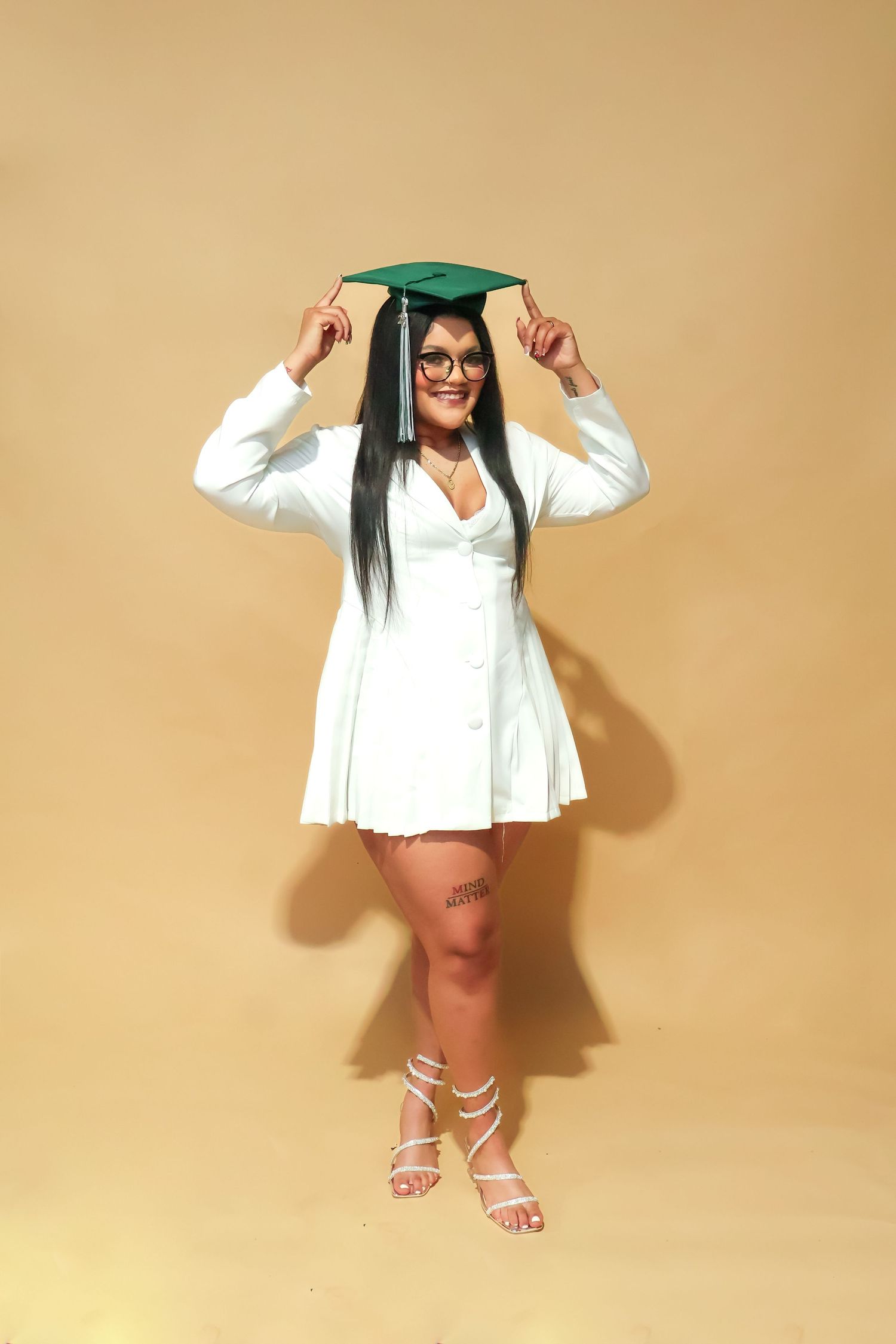 Smiling graduate in white dress holds green cap overhead on warm golden background.
