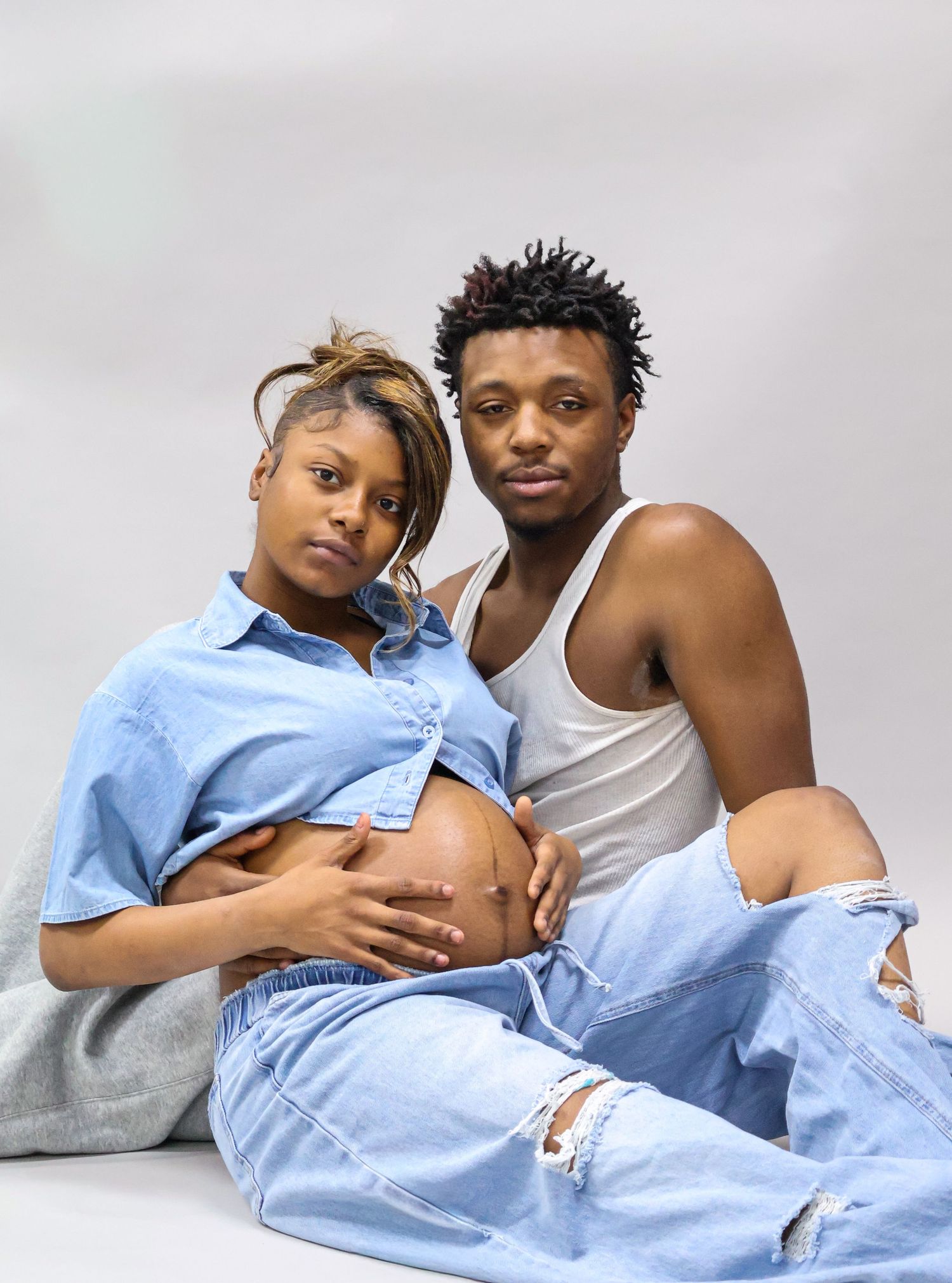 A pregnant couple poses together, woman in blue shirt showing baby bump, man in white tank and ripped jeans, studio shot.