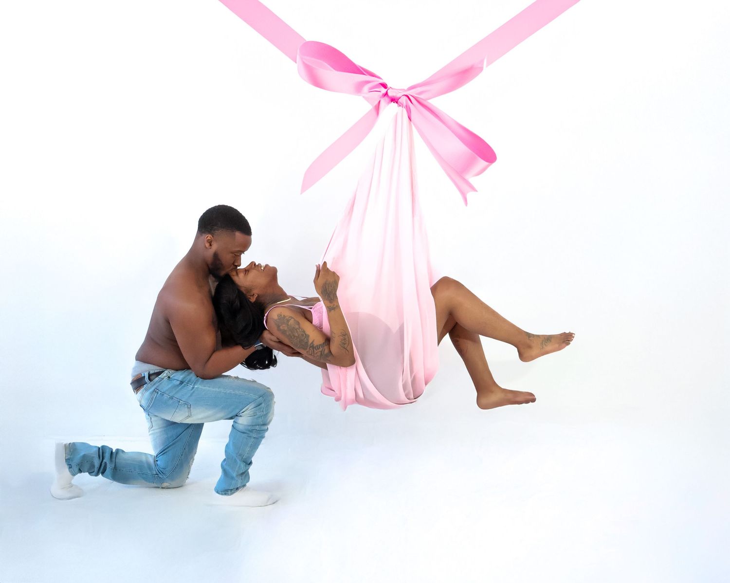 Couple poses romantically with woman in pink aerial silk swing and man kneeling beside her on white background.