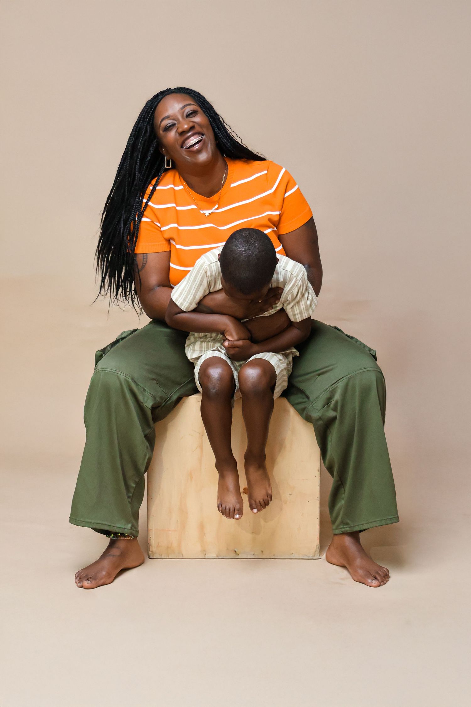 A joyful Black mother in an orange striped shirt hugs her young son on a wooden box against a beige background.