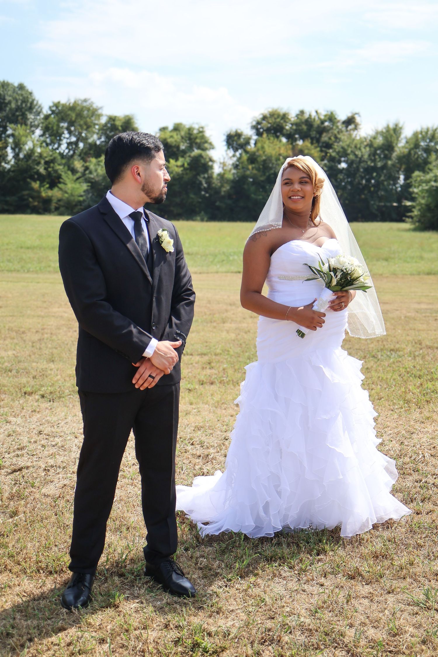 A bride in a white ruffled gown and veil holds a bouquet while standing with a groom in a black suit outdoors.