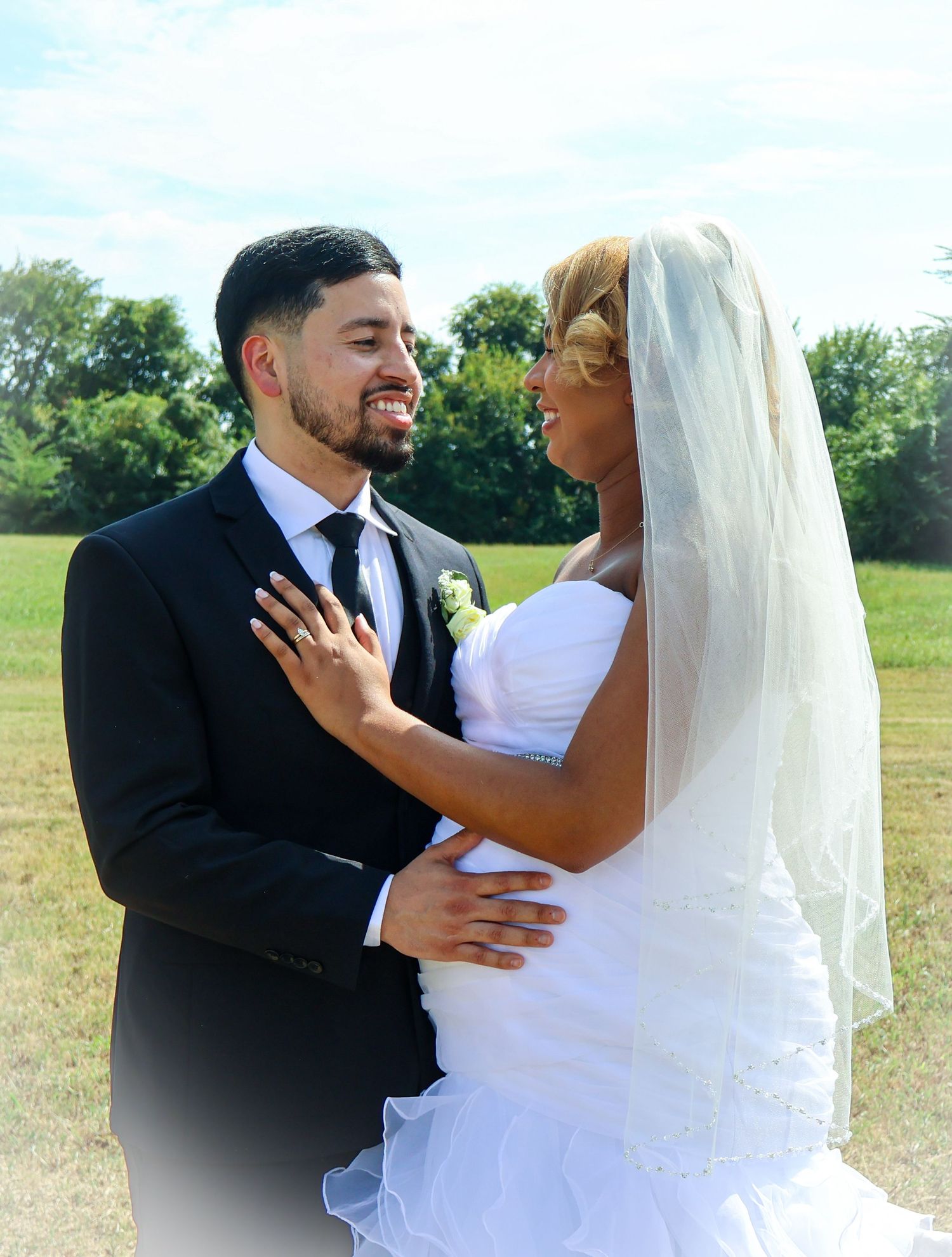 Smiling groom in black suit embracing bride in white wedding dress outdoors.