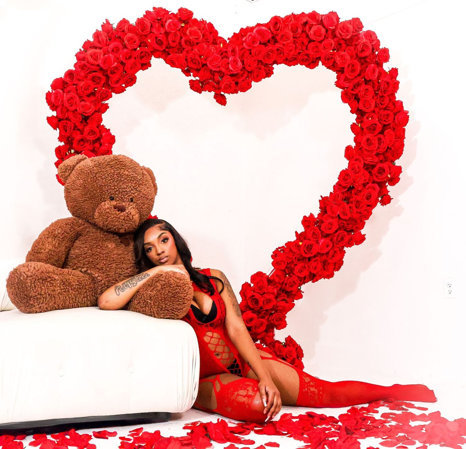 Woman in red lace outfit lounging with teddy bear near red rose heart backdrop.