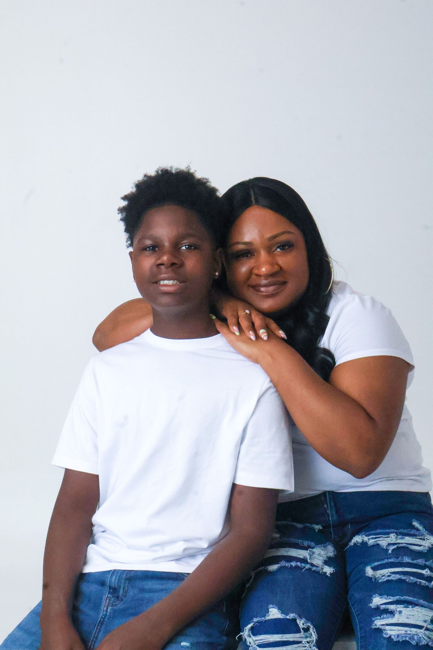 A mother and son wearing matching white tees and jeans pose together against a white background.