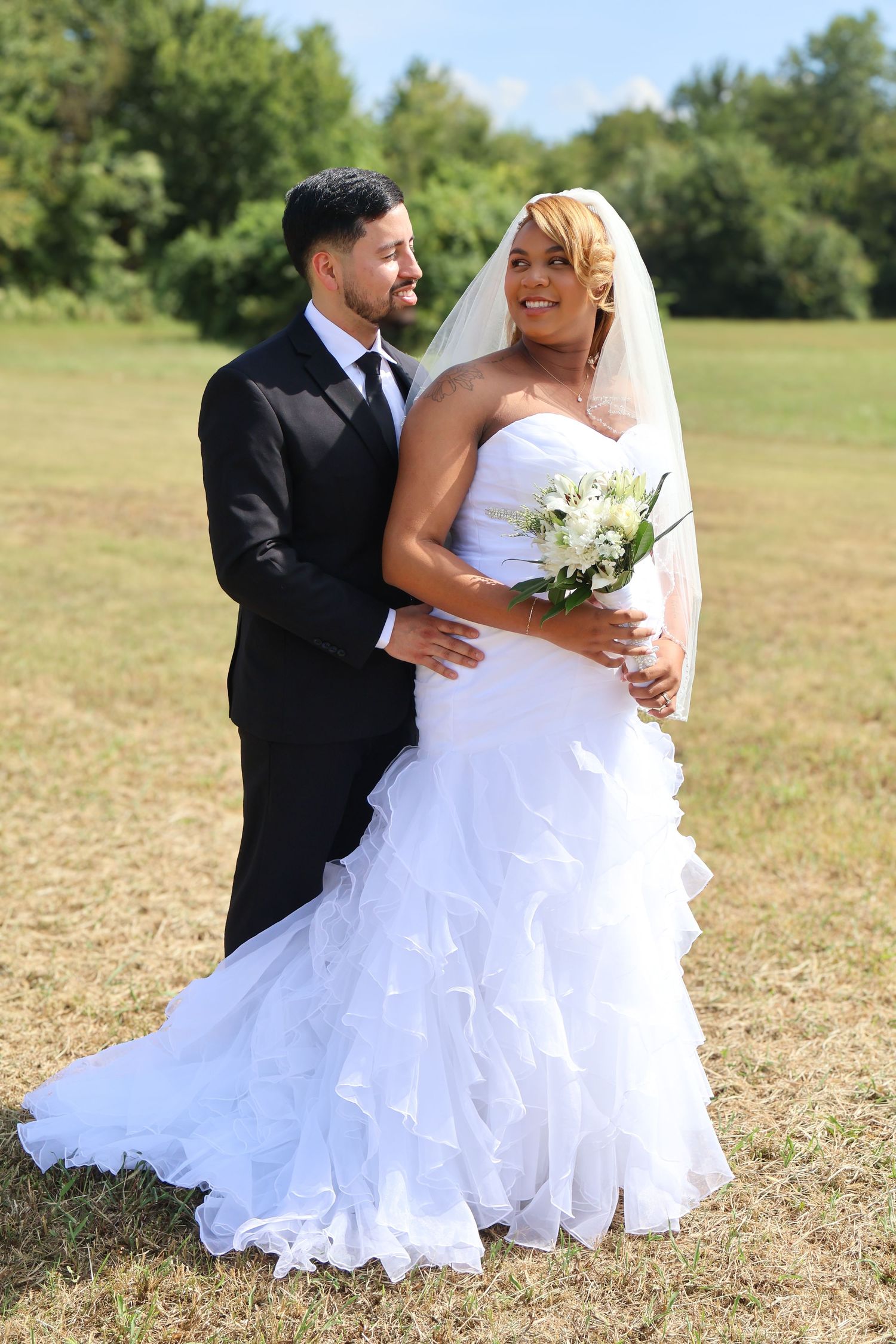 Bride in ruffled white gown holds bouquet while groom in black suit embraces her outdoors.