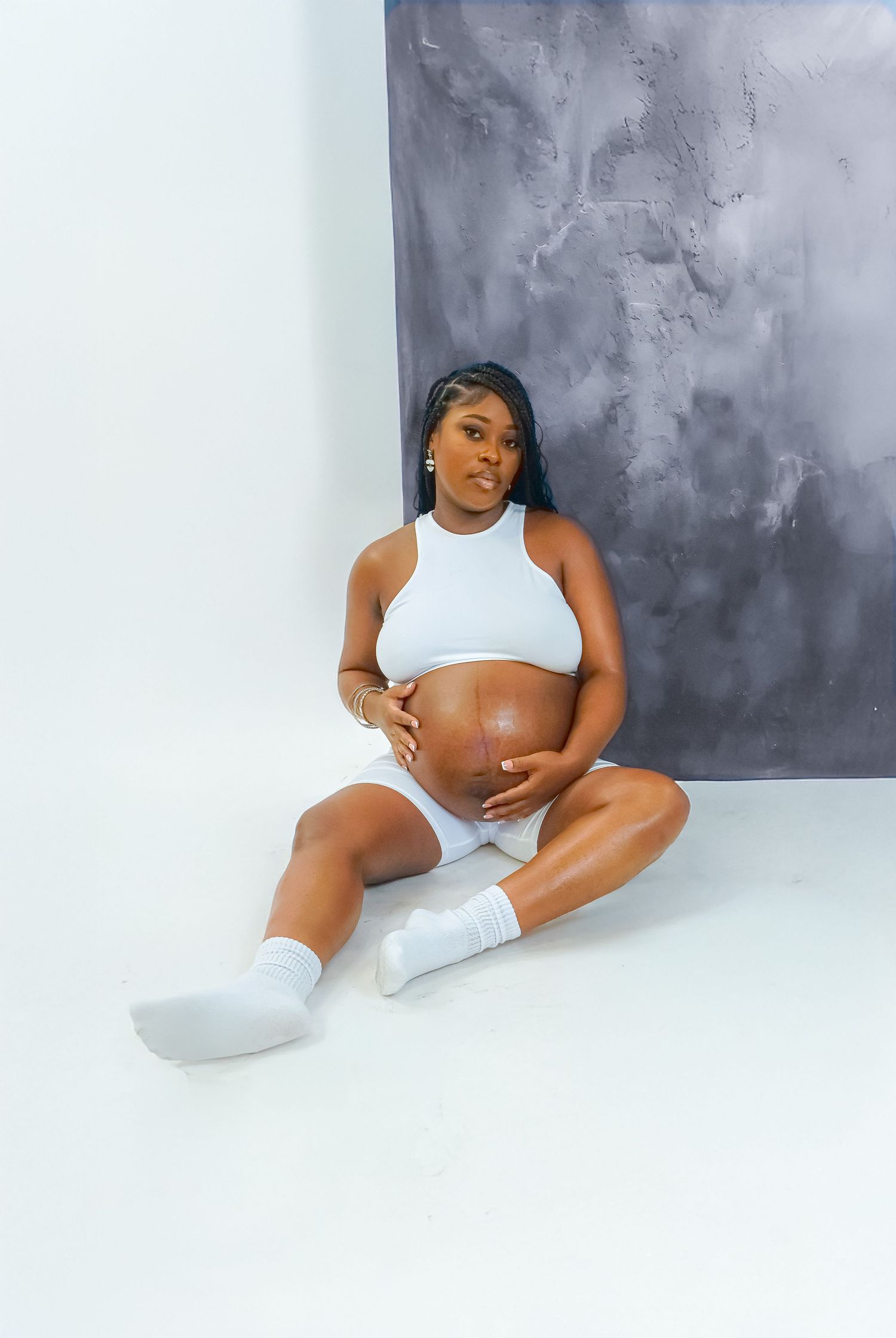 Pregnant woman in white crop top and shorts sitting on floor cradling baby bump during maternity photo shoot.