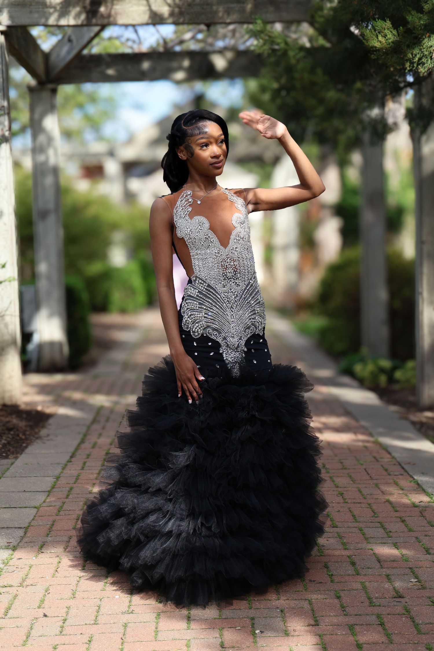 Young woman in elegant black mermaid gown with silver beading poses on brick path in lush garden setting.
