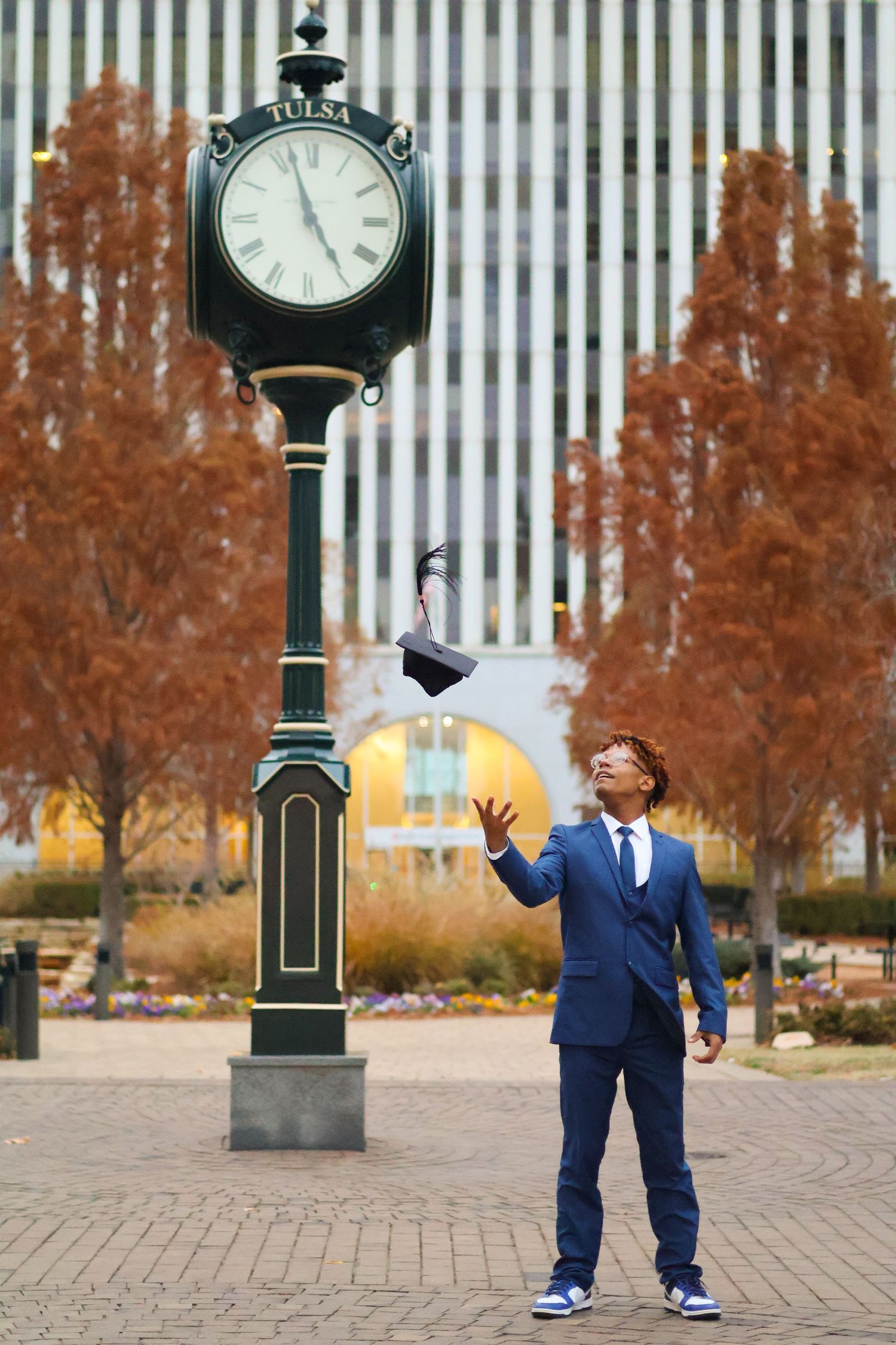 A young man in a blue suit tosses his graduation cap near a Tulsa street clock, celebrating his achievement outdoors.
