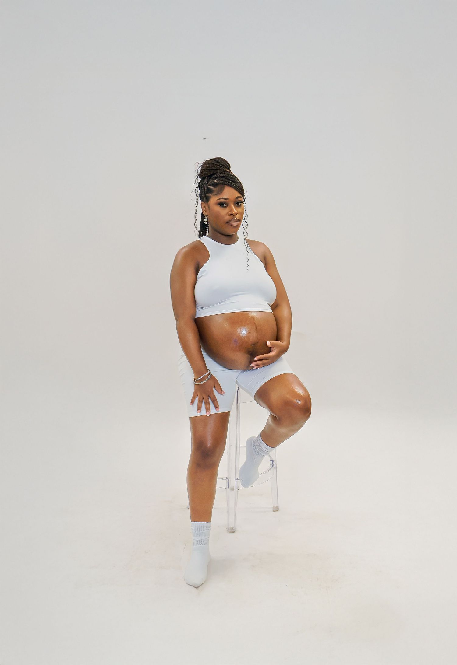 Pregnant woman in white crop top and shorts sits on clear chair, cradling baby bump in a studio setting.