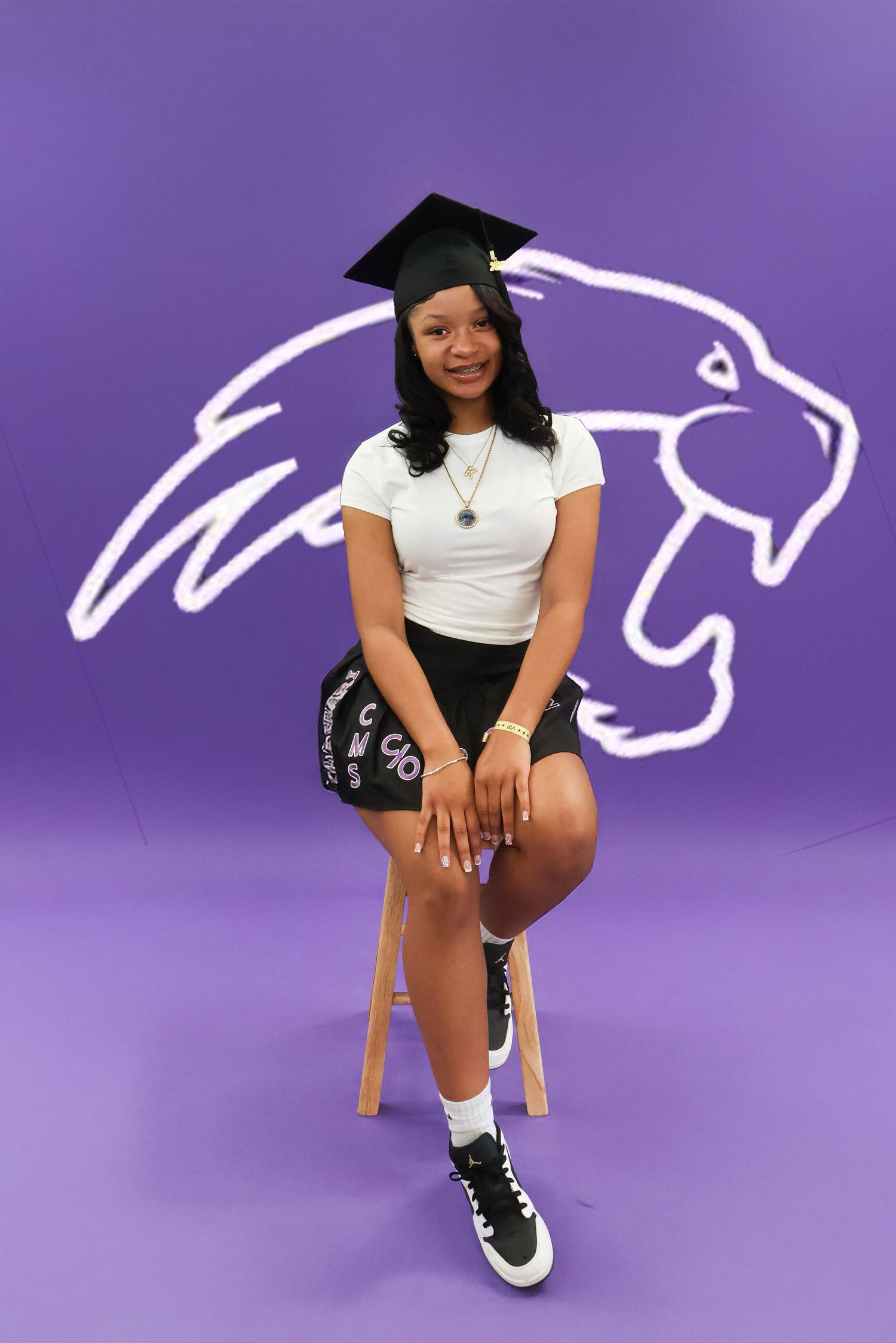 Young woman in graduation cap and black skirt sits before purple panther mascot backdrop.