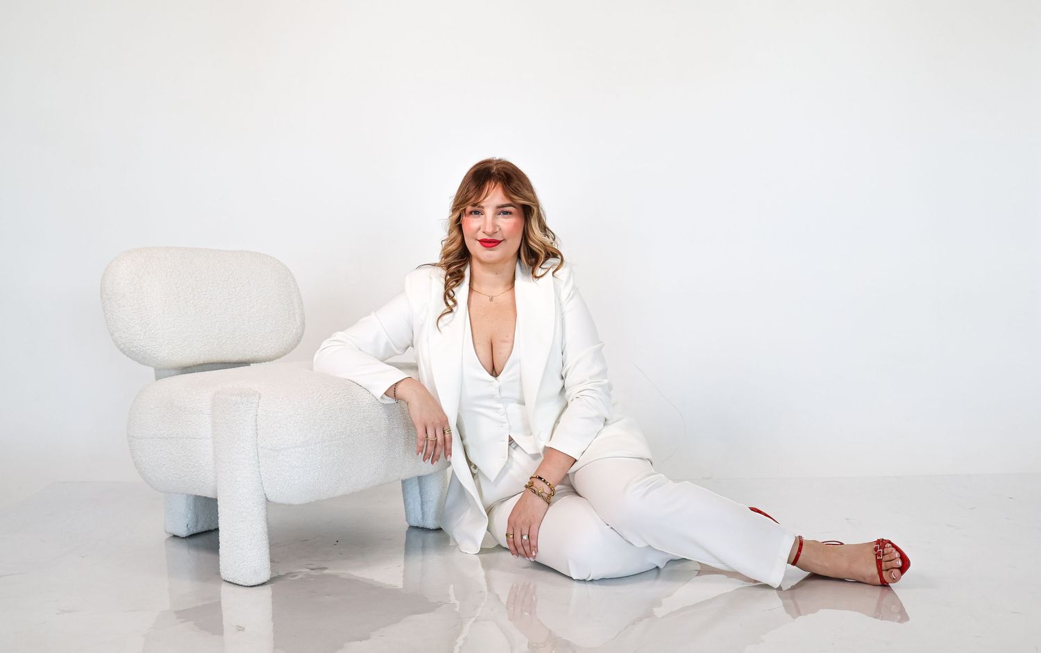 Woman in white blazer and pants sitting on floor beside modern white chair in minimalist studio.