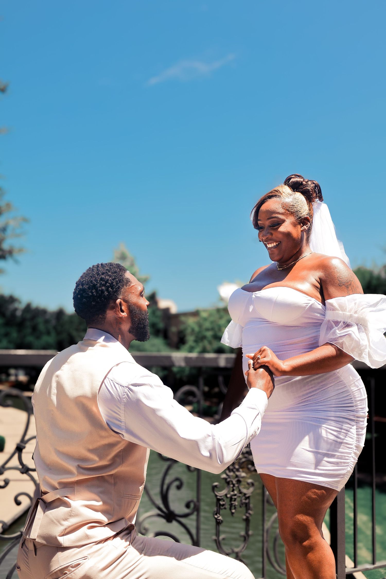 Smiling bride in white mini dress and veil holding hands with groom on sunny outdoor balcony.