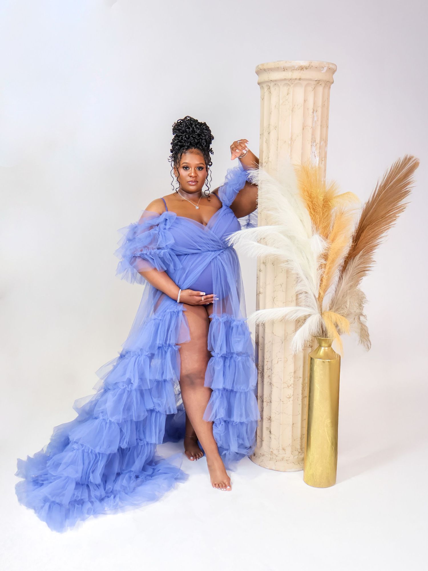 Pregnant woman in blue ruffled tulle maternity gown posing beside a decorative column with pampas grass.