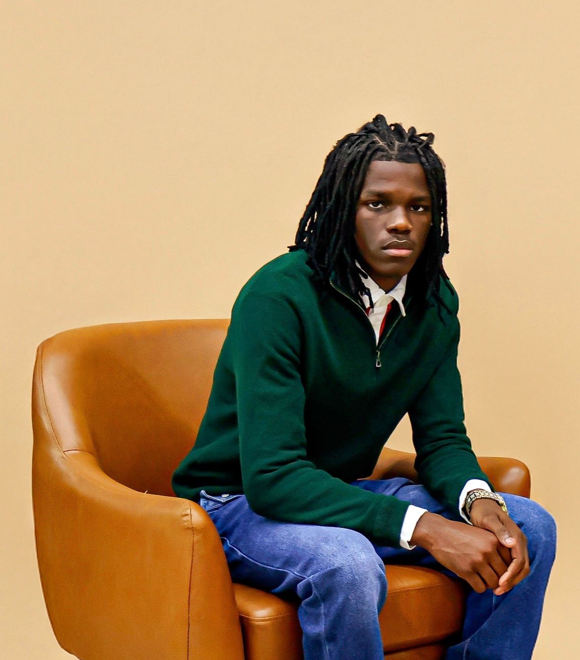 Young man with long braids sits in leather chair wearing green sweater and jeans.