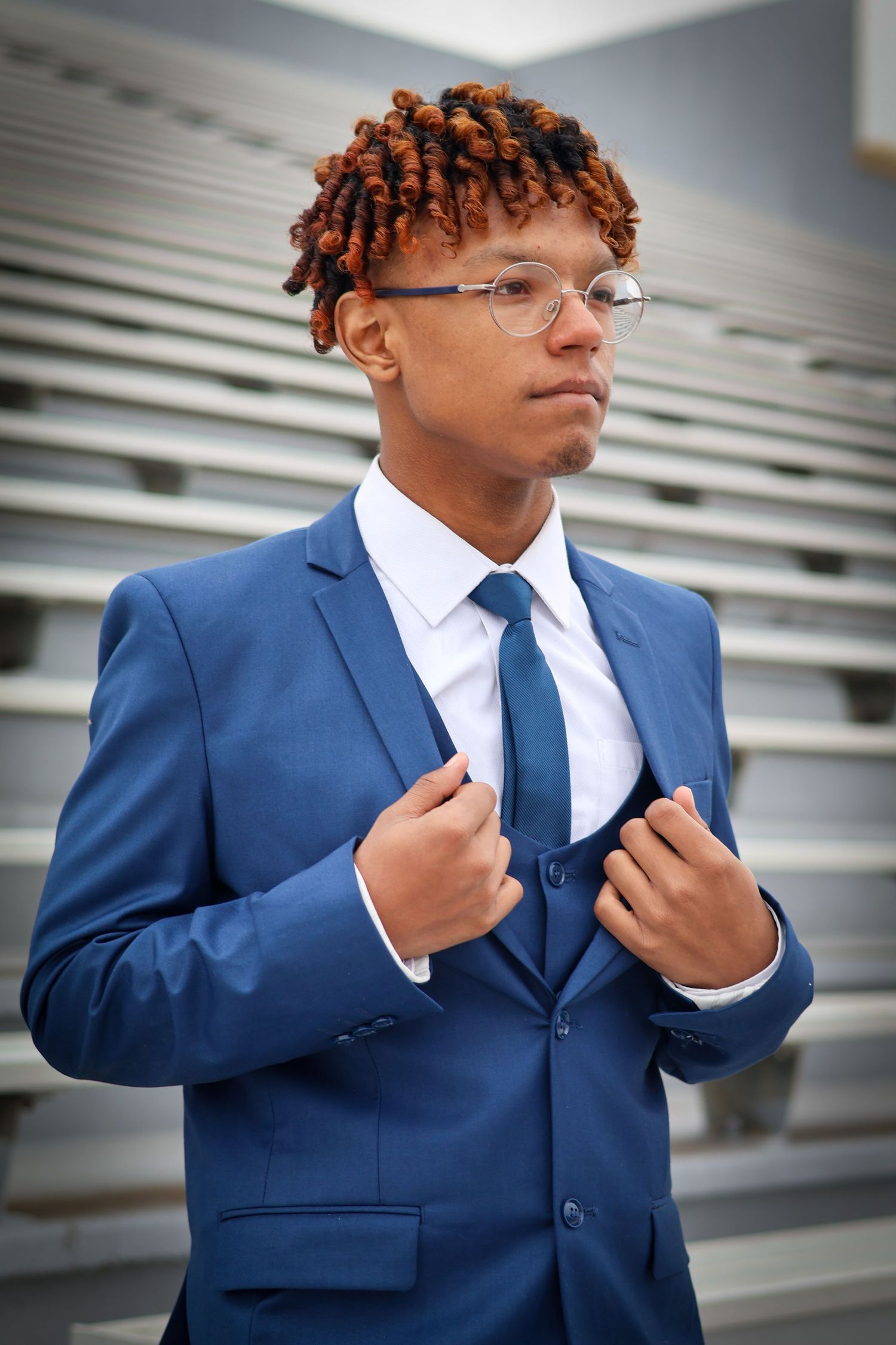 Young Black man with red locs in blue suit and glasses adjusts jacket near stadium seating.