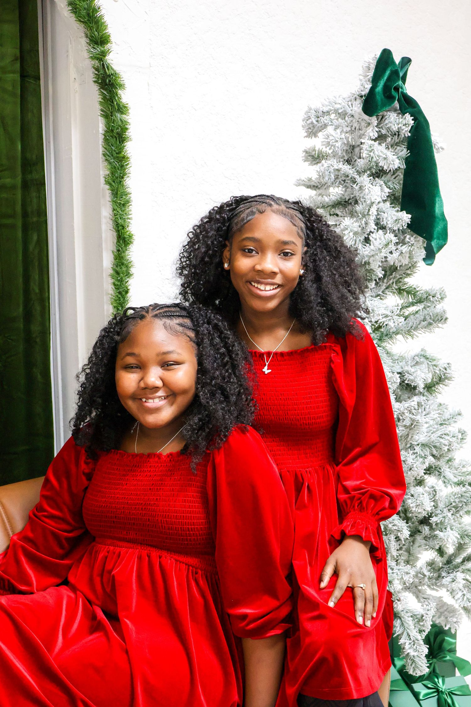 Two young girls in matching red velvet dresses smiling beside a flocked Christmas tree.
