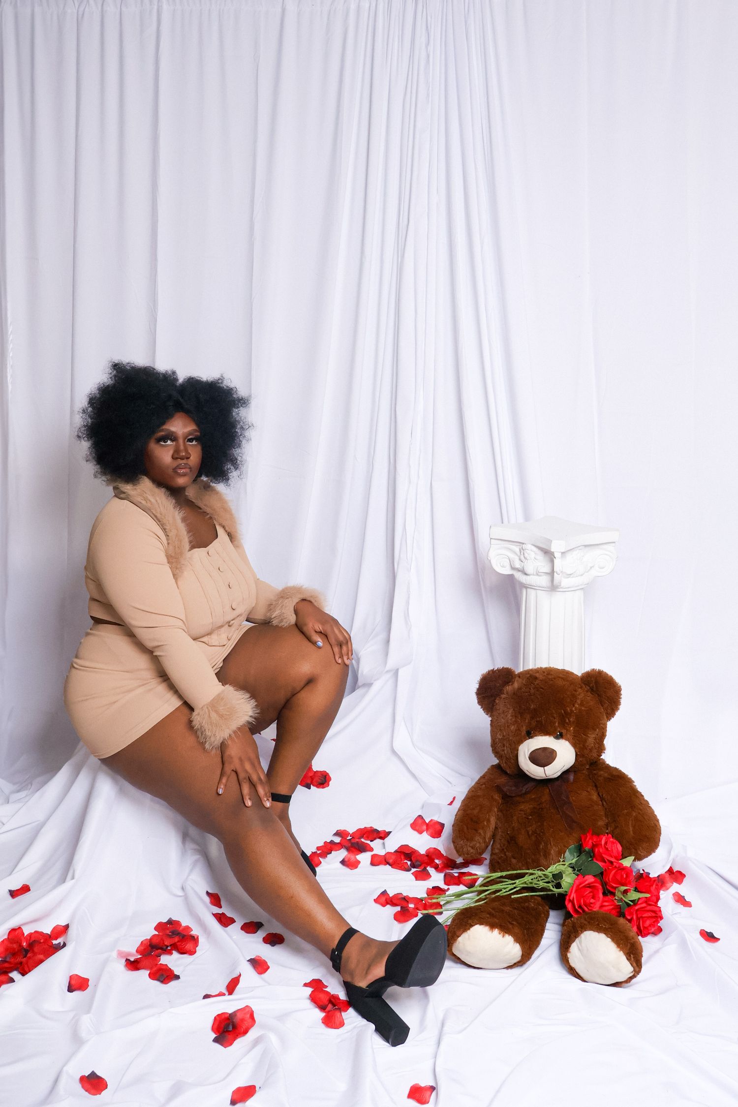 Plus-size woman in beige dress sitting among rose petals with teddy bear holding roses on white backdrop.