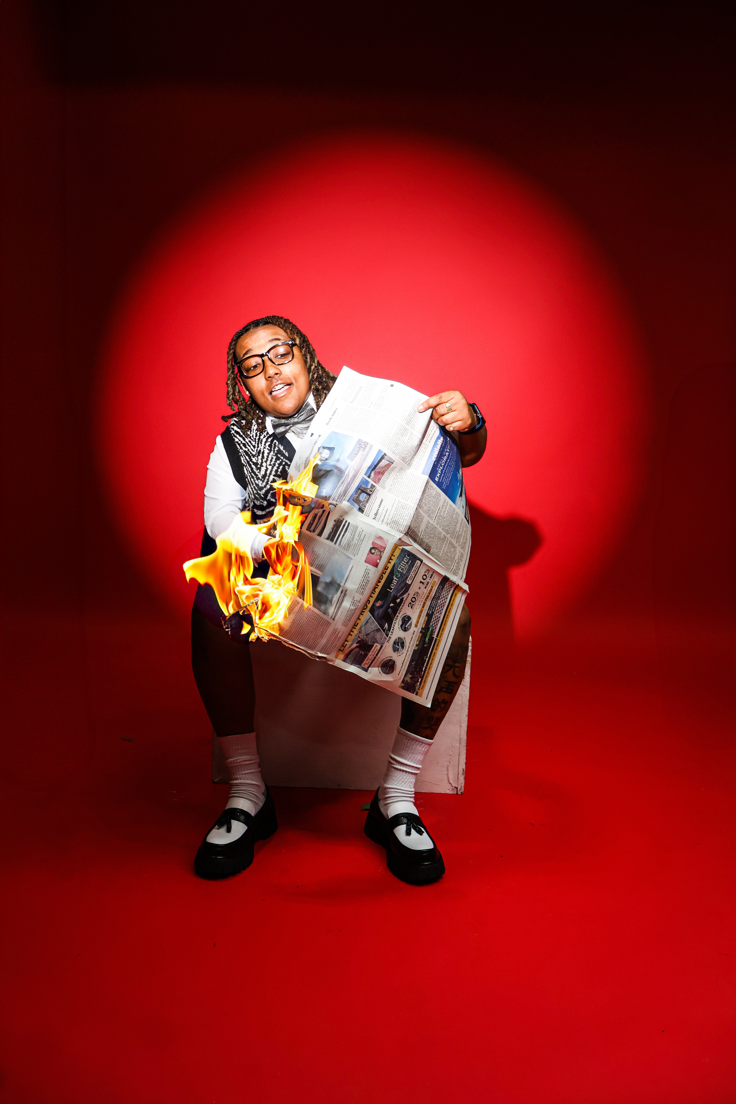Person holding a burning newspaper on a dramatic red spotlight background wearing glasses and casual outfit.
