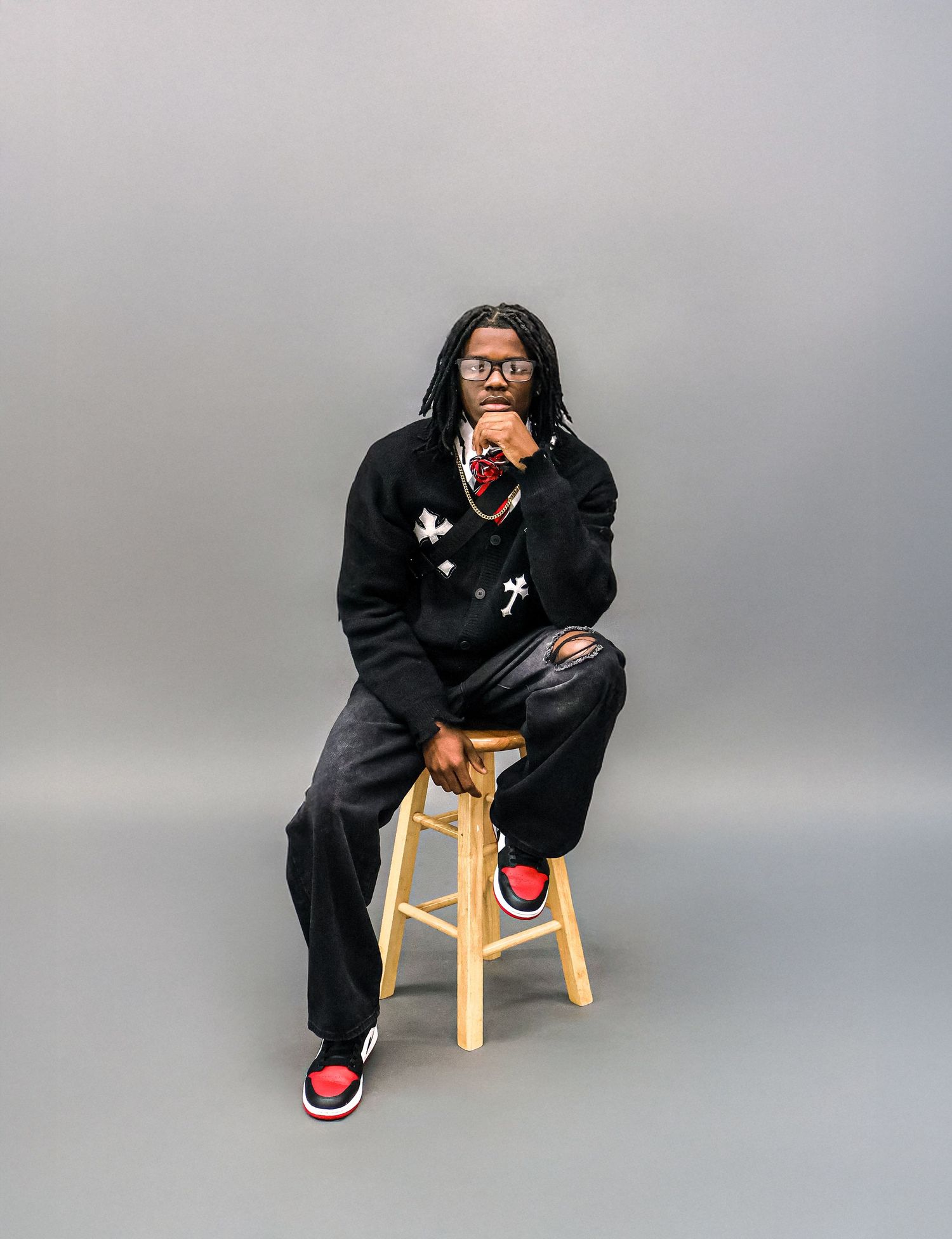 Young man with braids in black outfit and red Jordans sits on wooden stool.