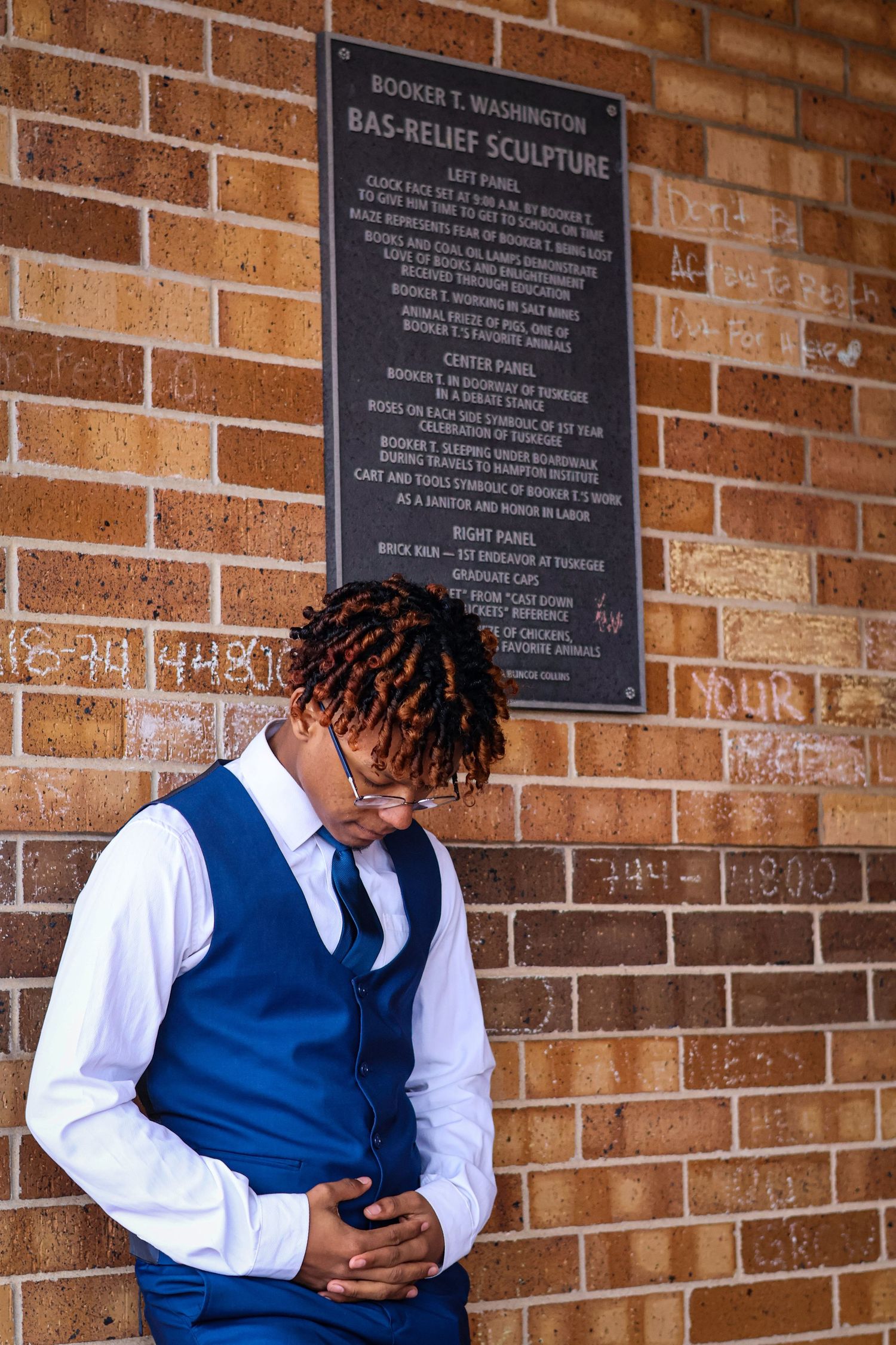 Young Black man in blue vest bows head near Booker T. Washington Bas-Relief Sculpture plaque.