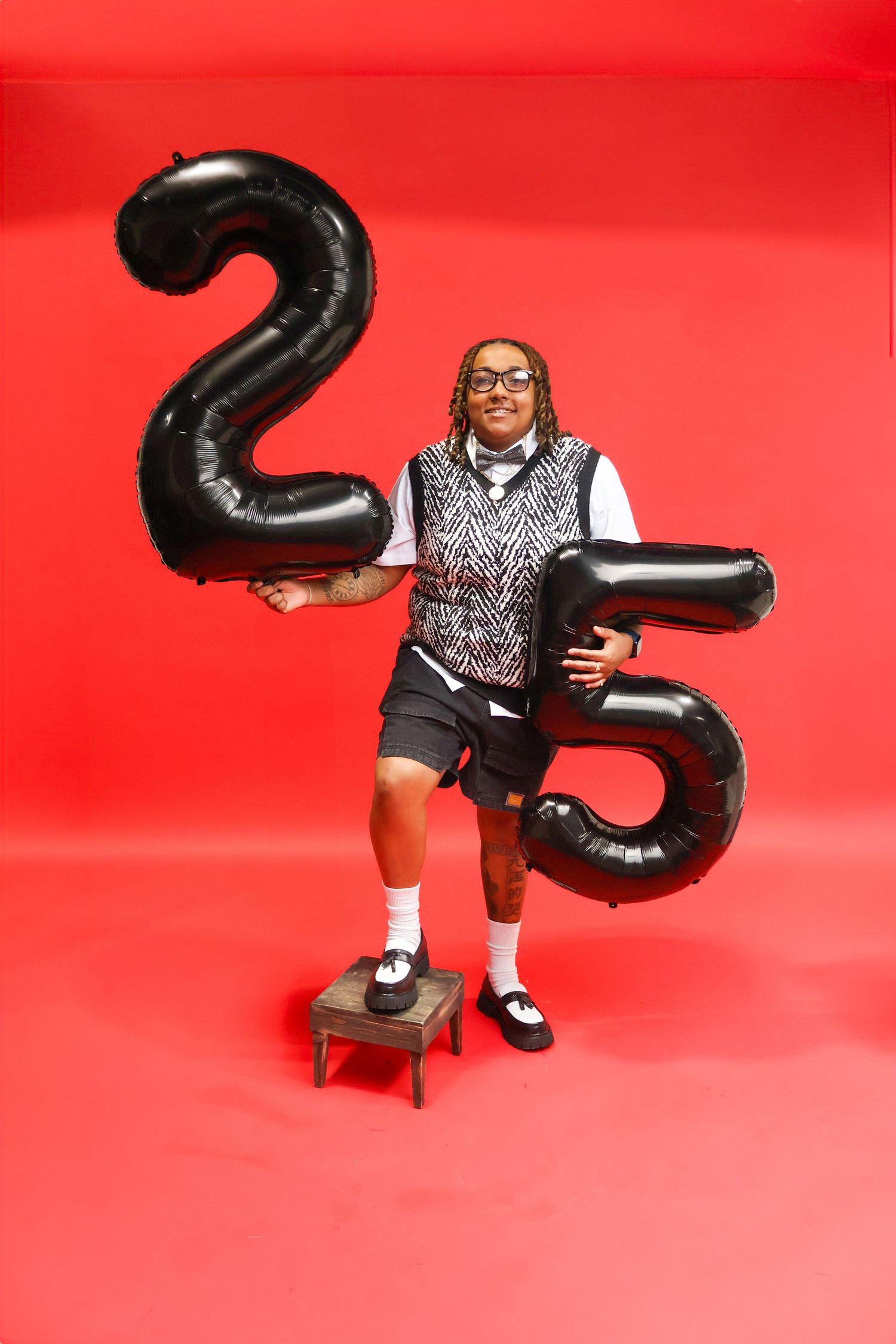 Person standing on stool holding large black number 25 balloons on red background in nerd costume.
