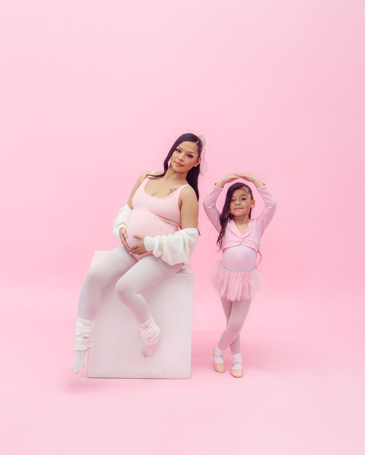 Pregnant woman in pink ballet outfit poses with young girl in pink tutu on pink background.