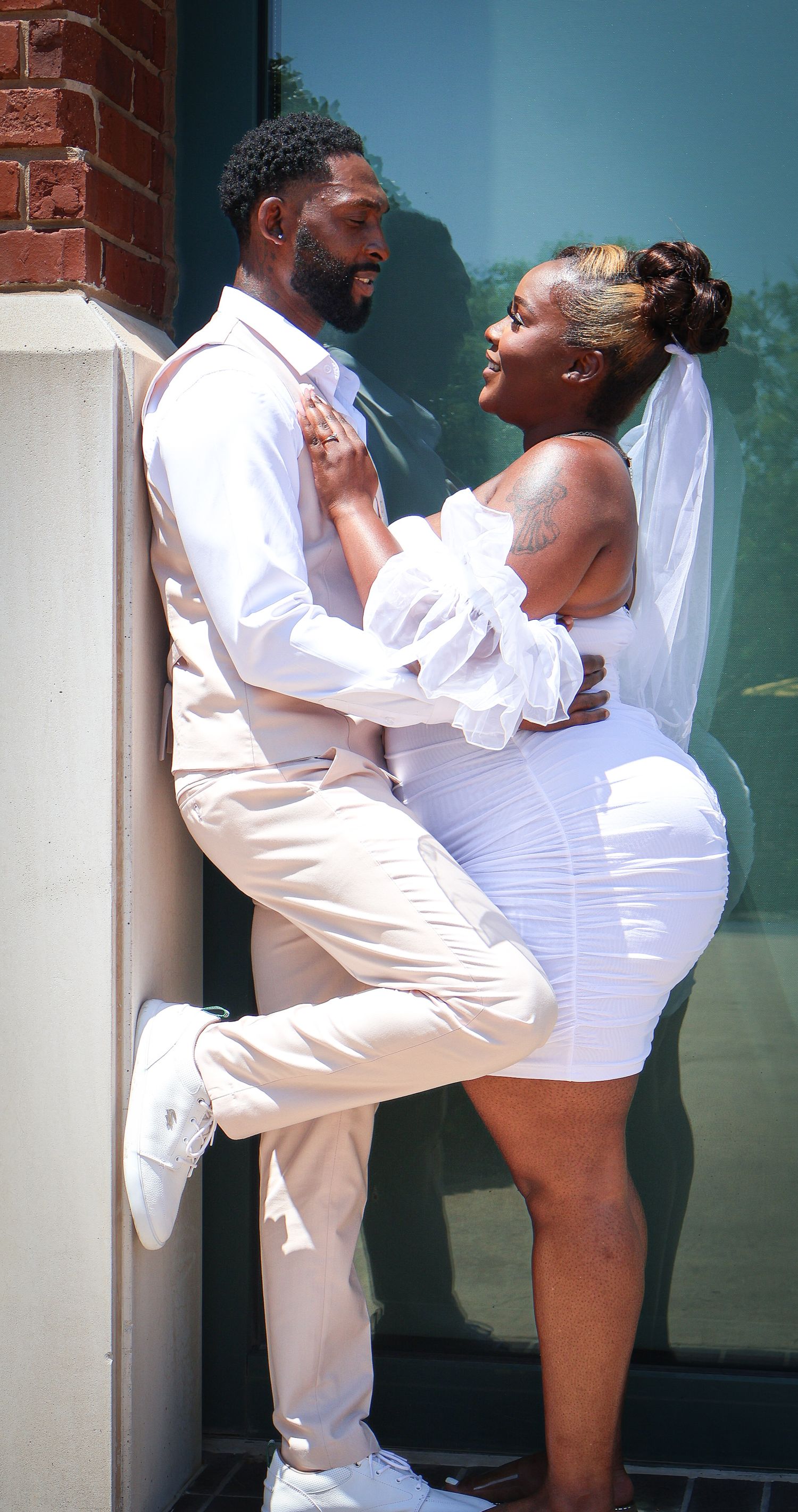 Bride and groom in white attire sharing romantic moment leaning against brick wall outdoors.