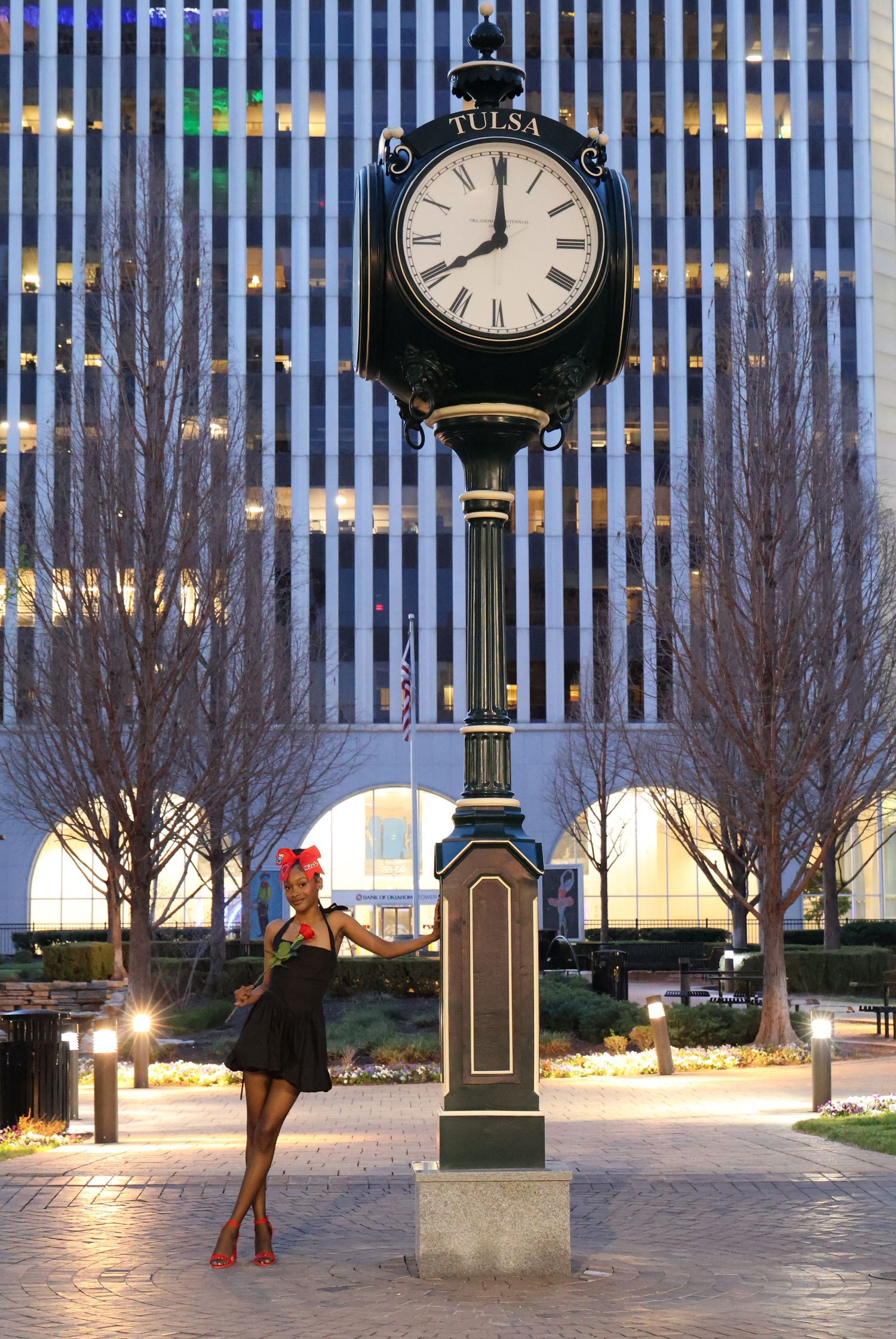Young woman in black dress posing beside a Tulsa street clock at night in an urban plaza.