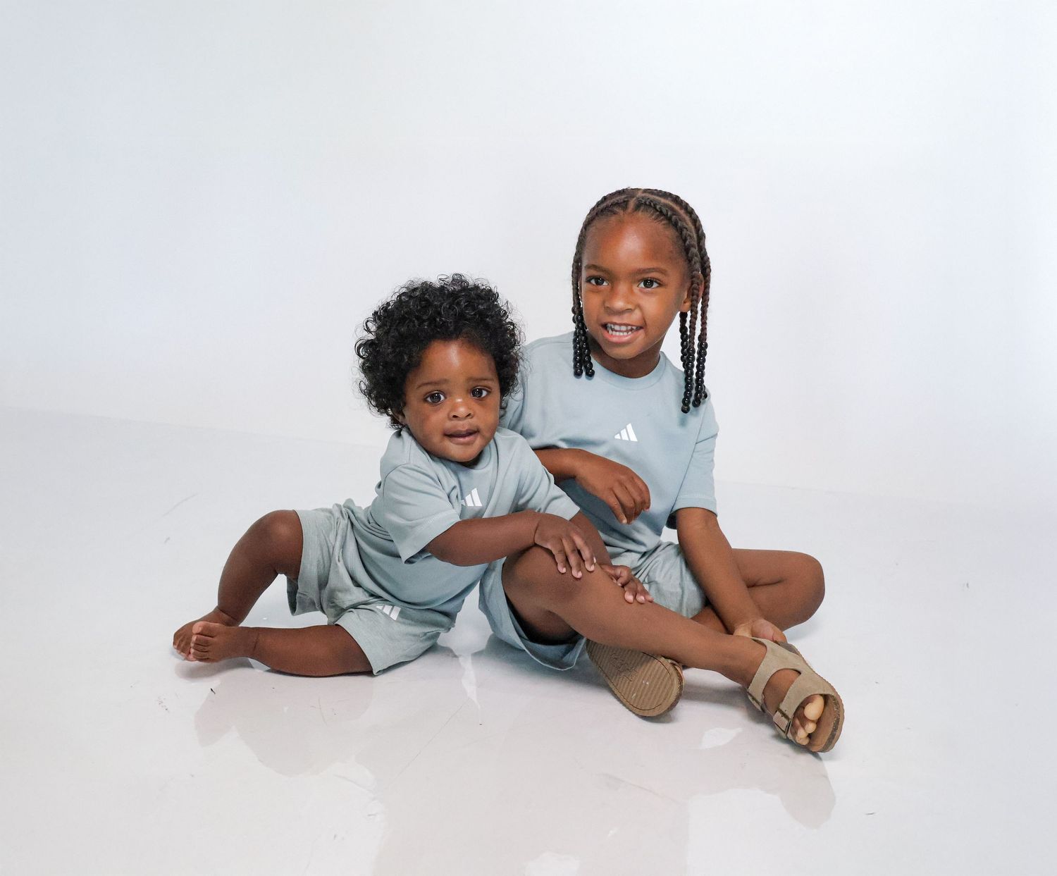 Two young children in matching light blue outfits sitting together and smiling against a white background.