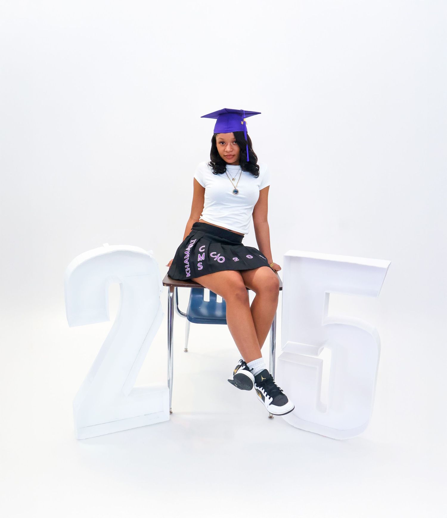 Graduate in purple cap and decorated skirt sits beside '25' props on white backdrop.