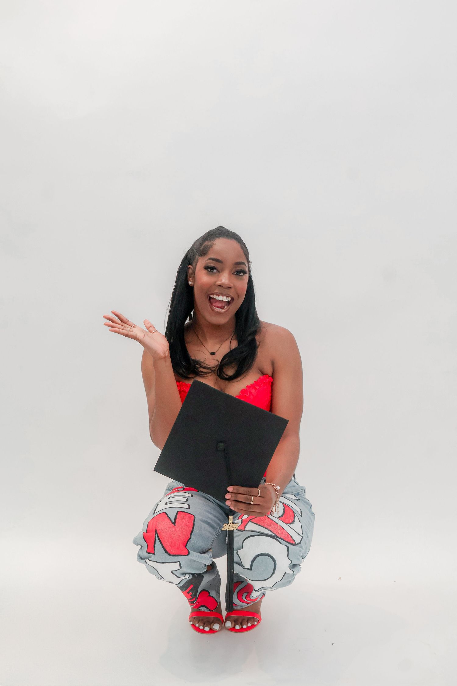 Excited graduate crouches holding black mortarboard cap in red top and graphic jeans.