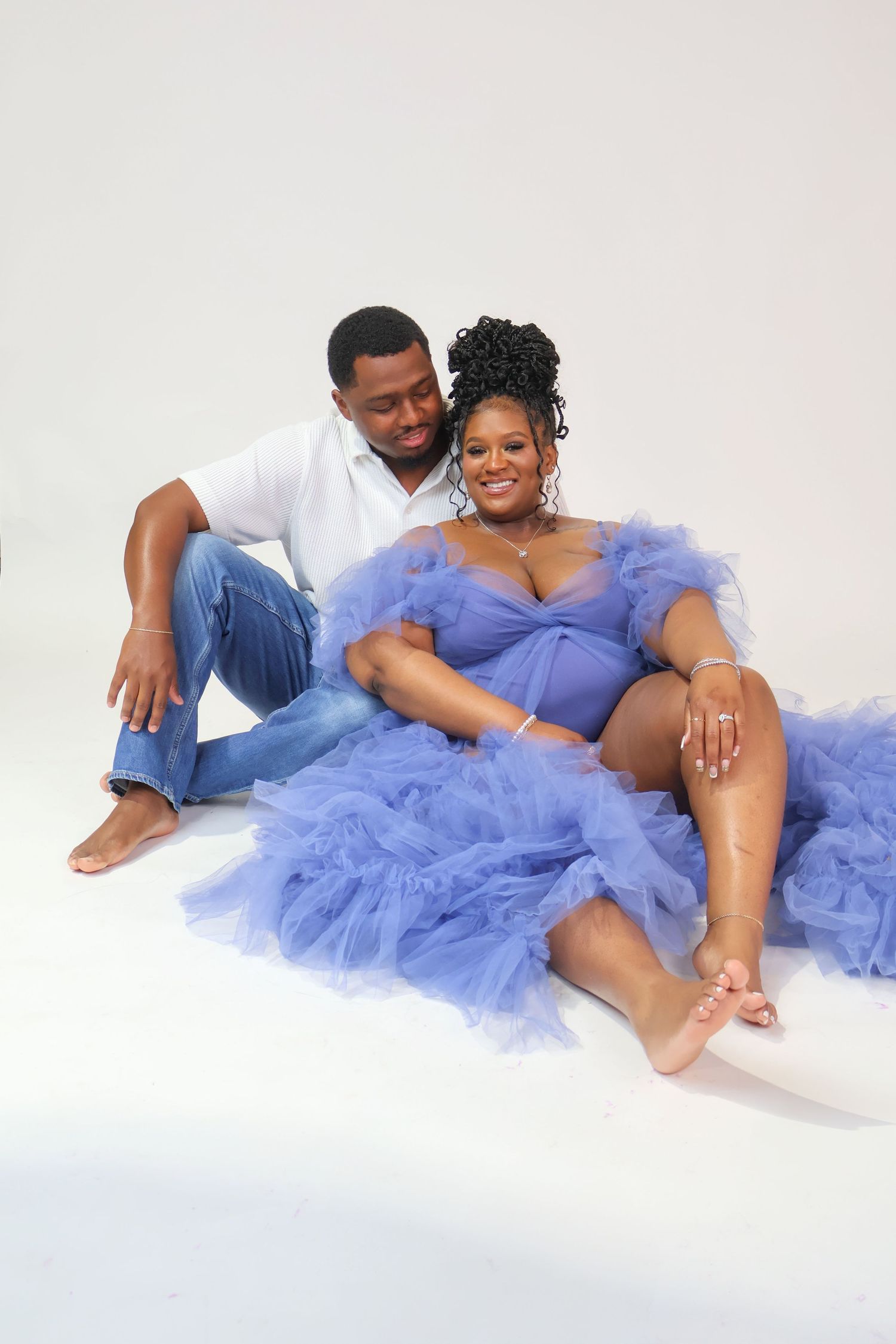 Couple poses for maternity photo shoot, woman in blue tulle gown, man in white shirt and jeans, white studio background.