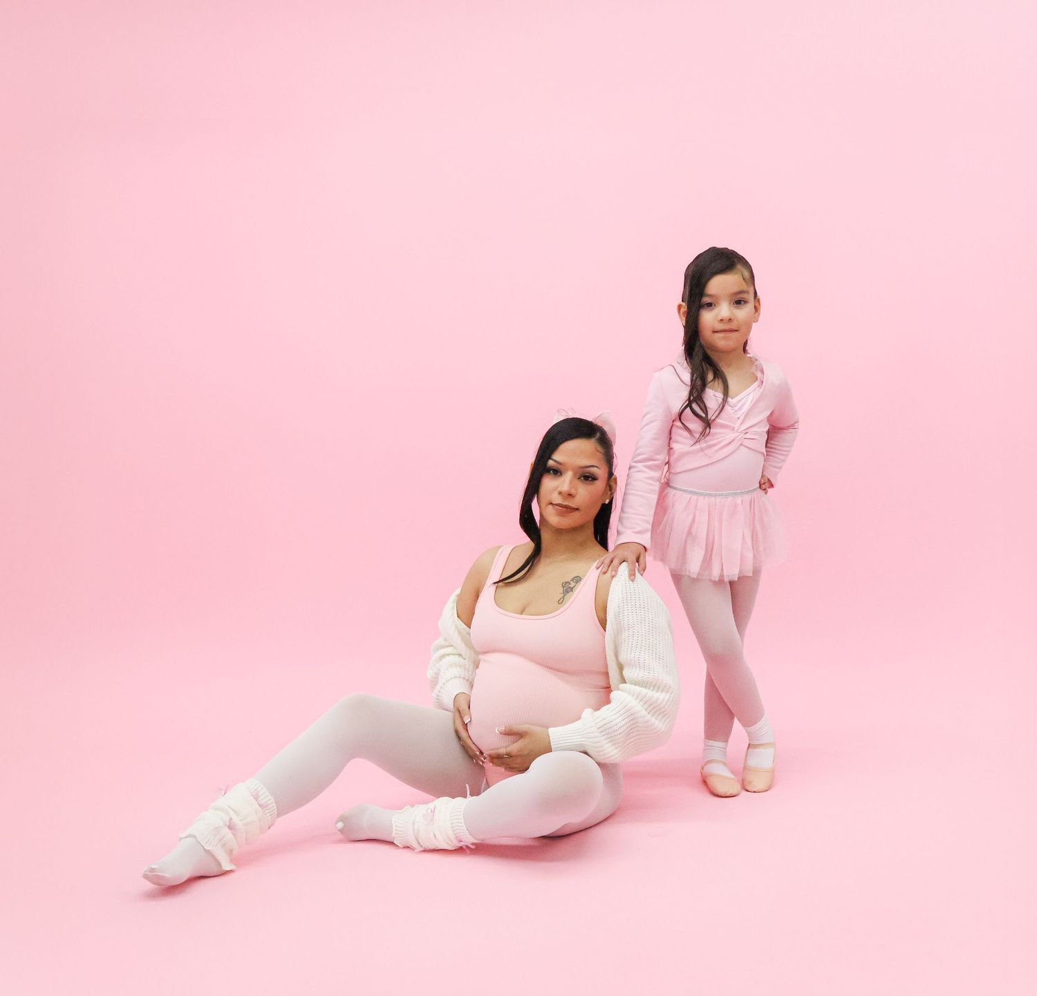Pregnant woman in white outfit sits beside a young girl in pink ballet attire against a pink background.