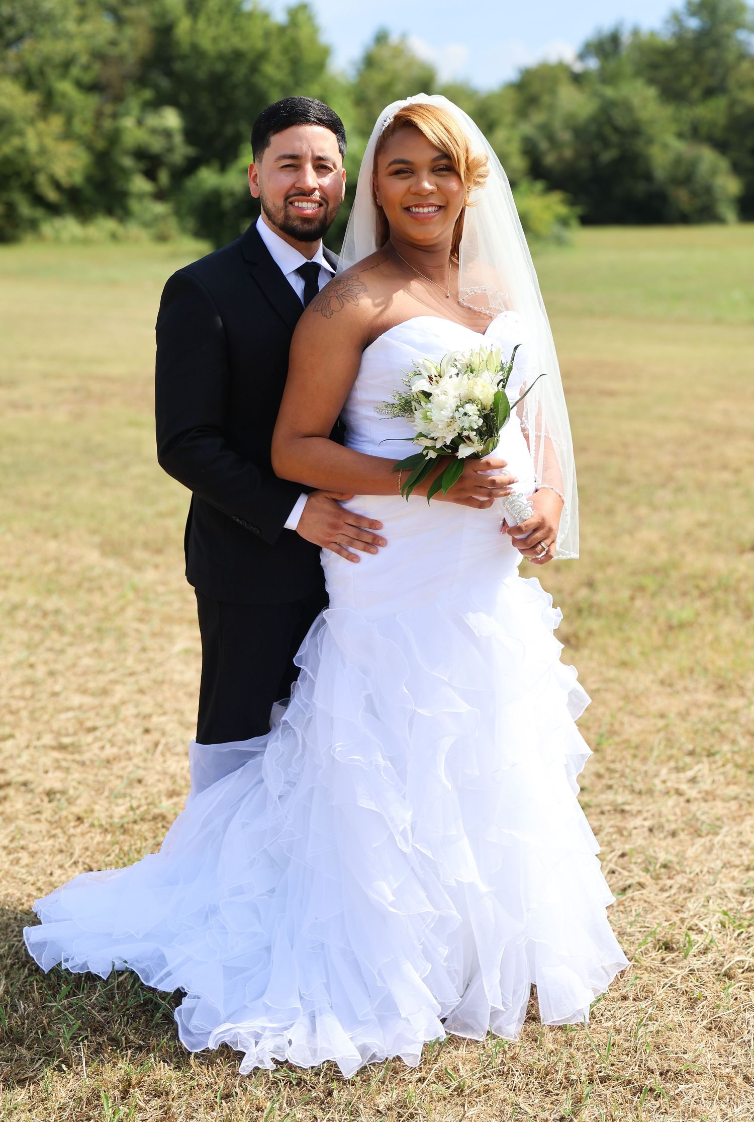 Bride in white ruffled gown holding bouquet with groom in black suit posing outdoors on a sunny wedding day.