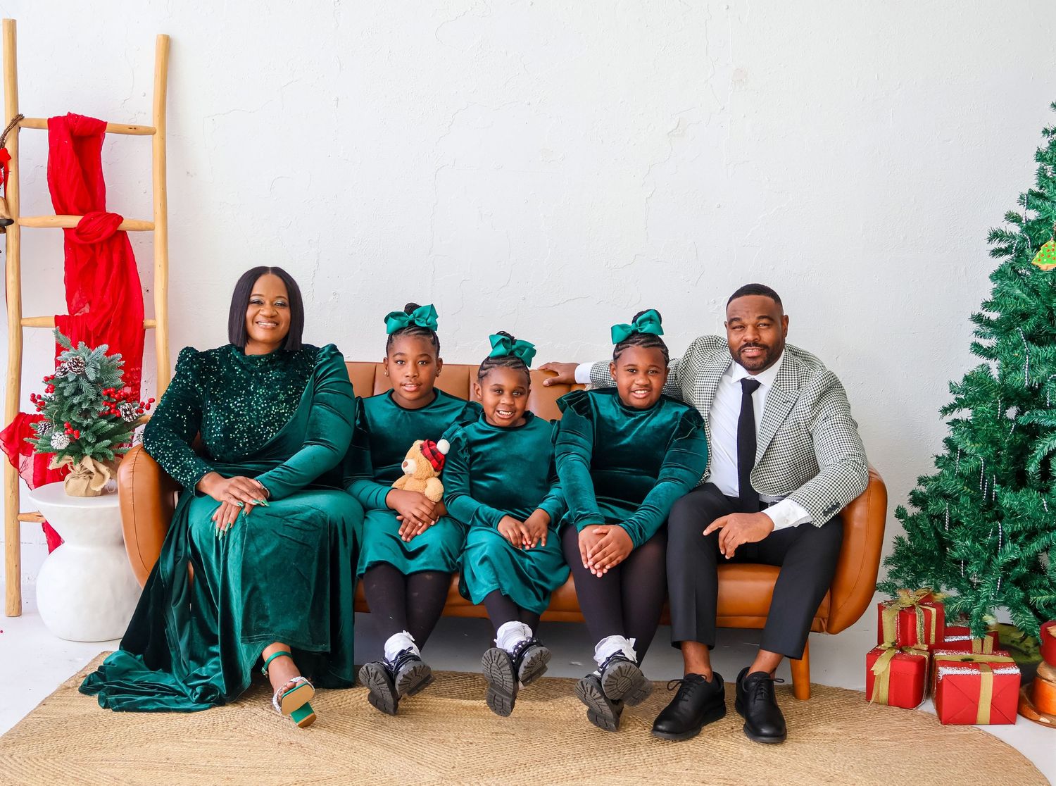 Family of five in matching green outfits posing on a couch with Christmas tree and gifts in a festive holiday photo.