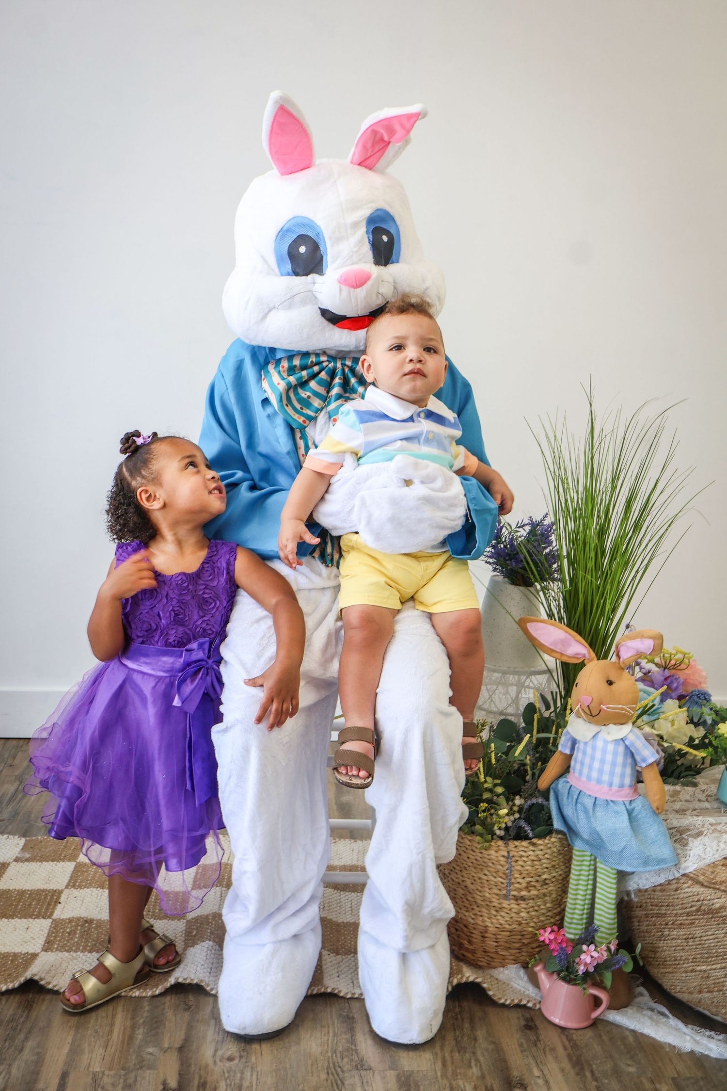 Toddler girl in purple dress and baby boy sitting with Easter Bunny mascot in spring photo session.