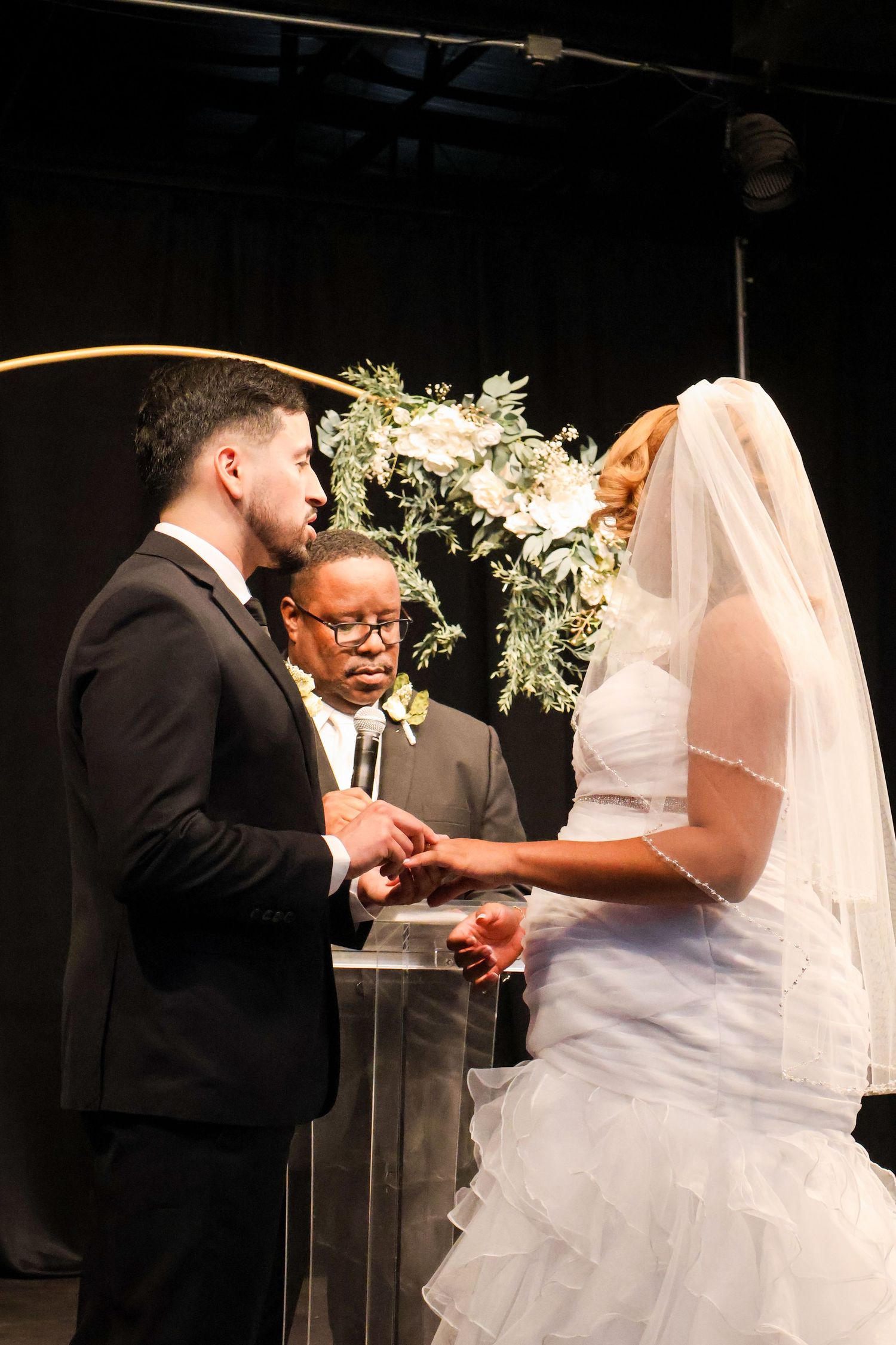 Groom holds brides hands during wedding ceremony as officiant reads vows at floral arch altar.