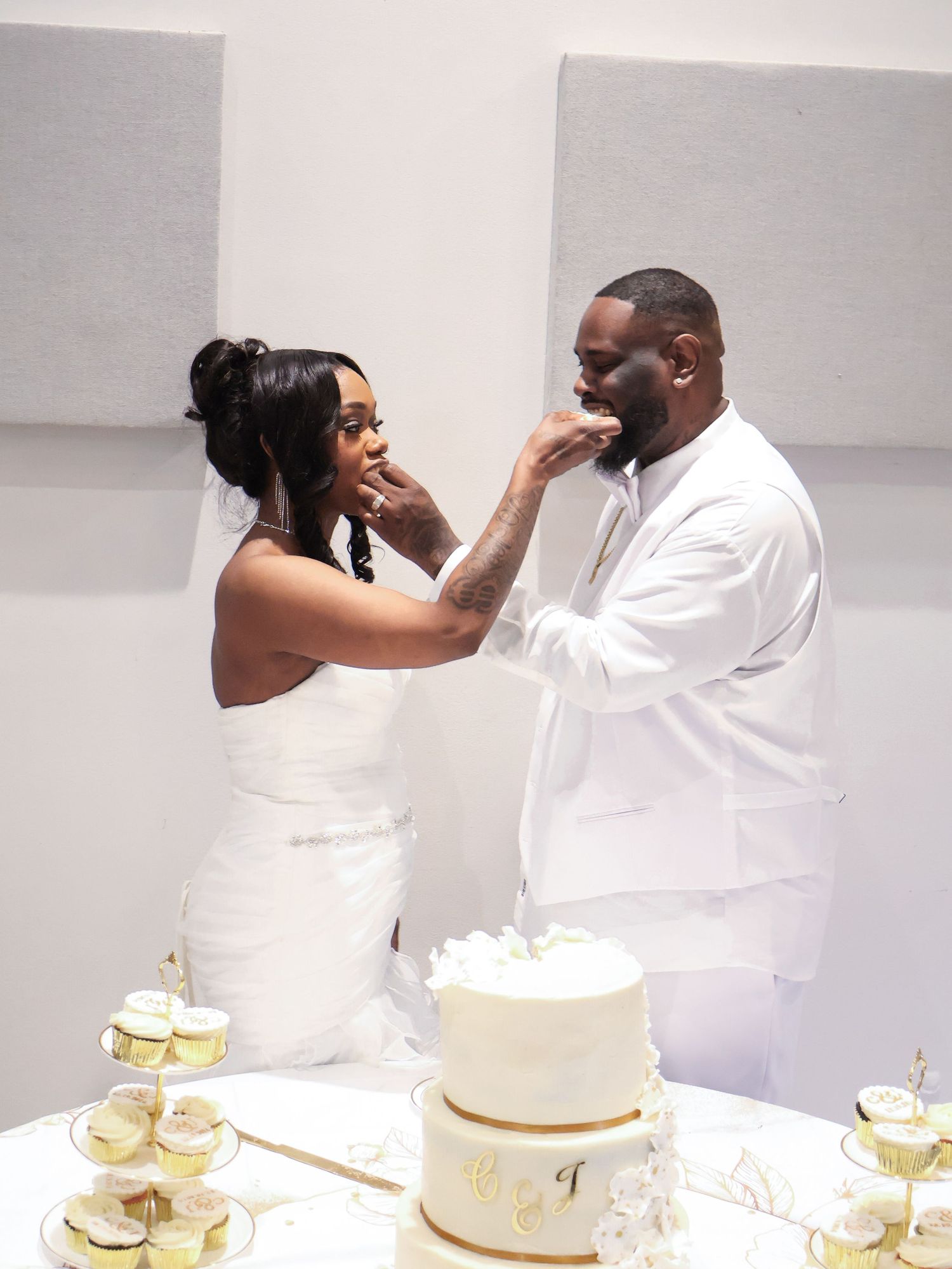 Newlyweds feeding each other wedding cake at elegant gold and white reception table.
