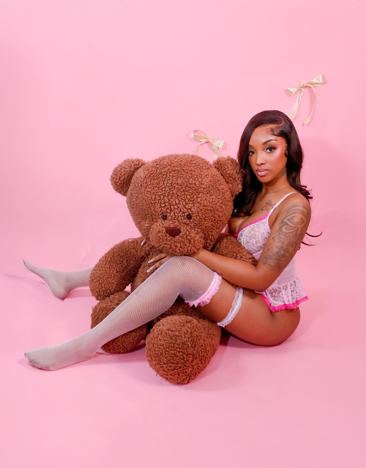 Woman in white lingerie posing with large brown teddy bear on pink background.