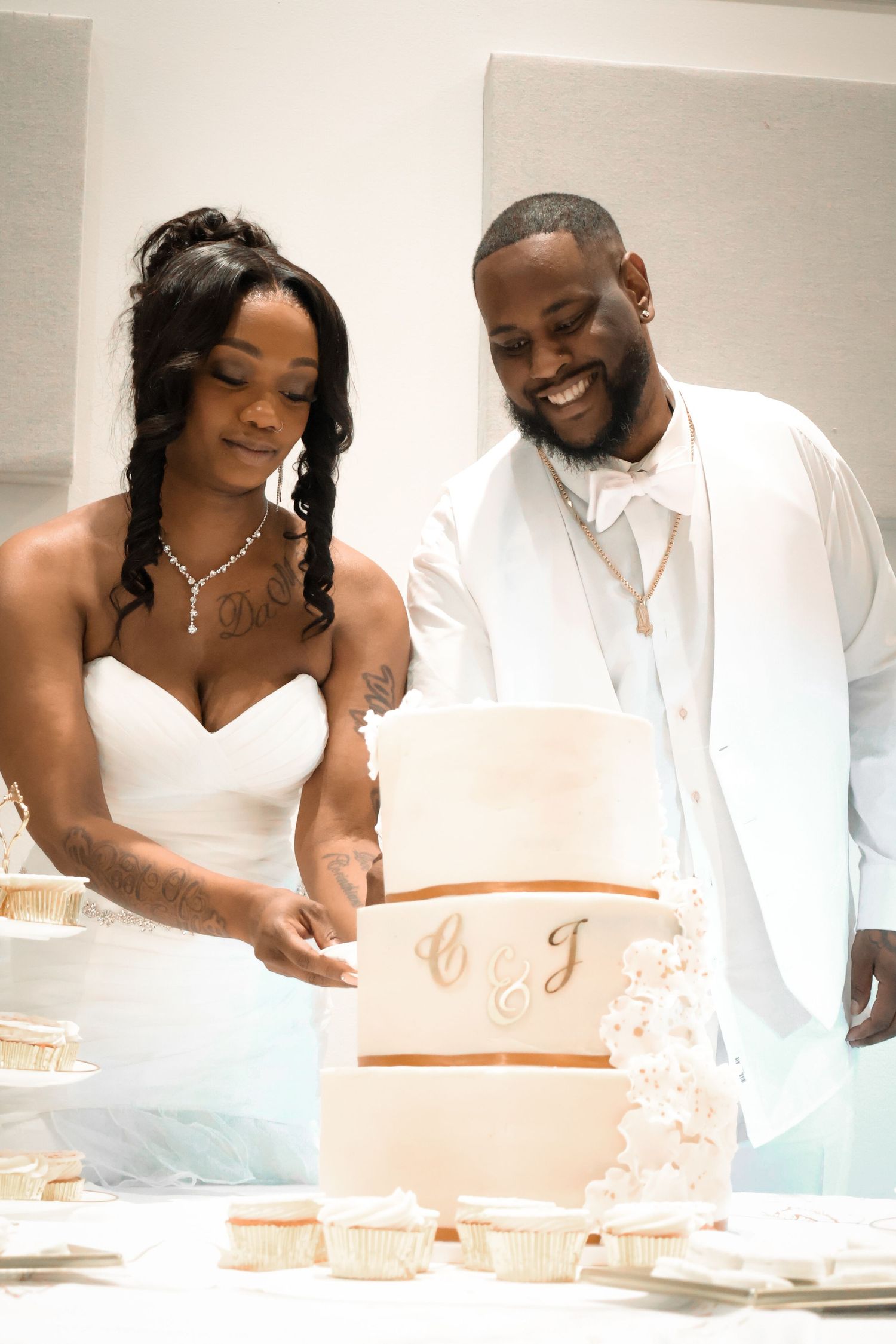 Bride and groom in white attire cut elegant three-tier wedding cake with gold monogram details.