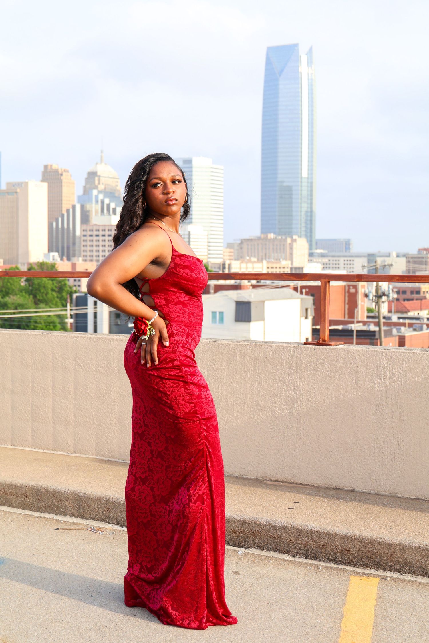 A woman in a red sequin gown poses on a rooftop with a city skyline behind her.