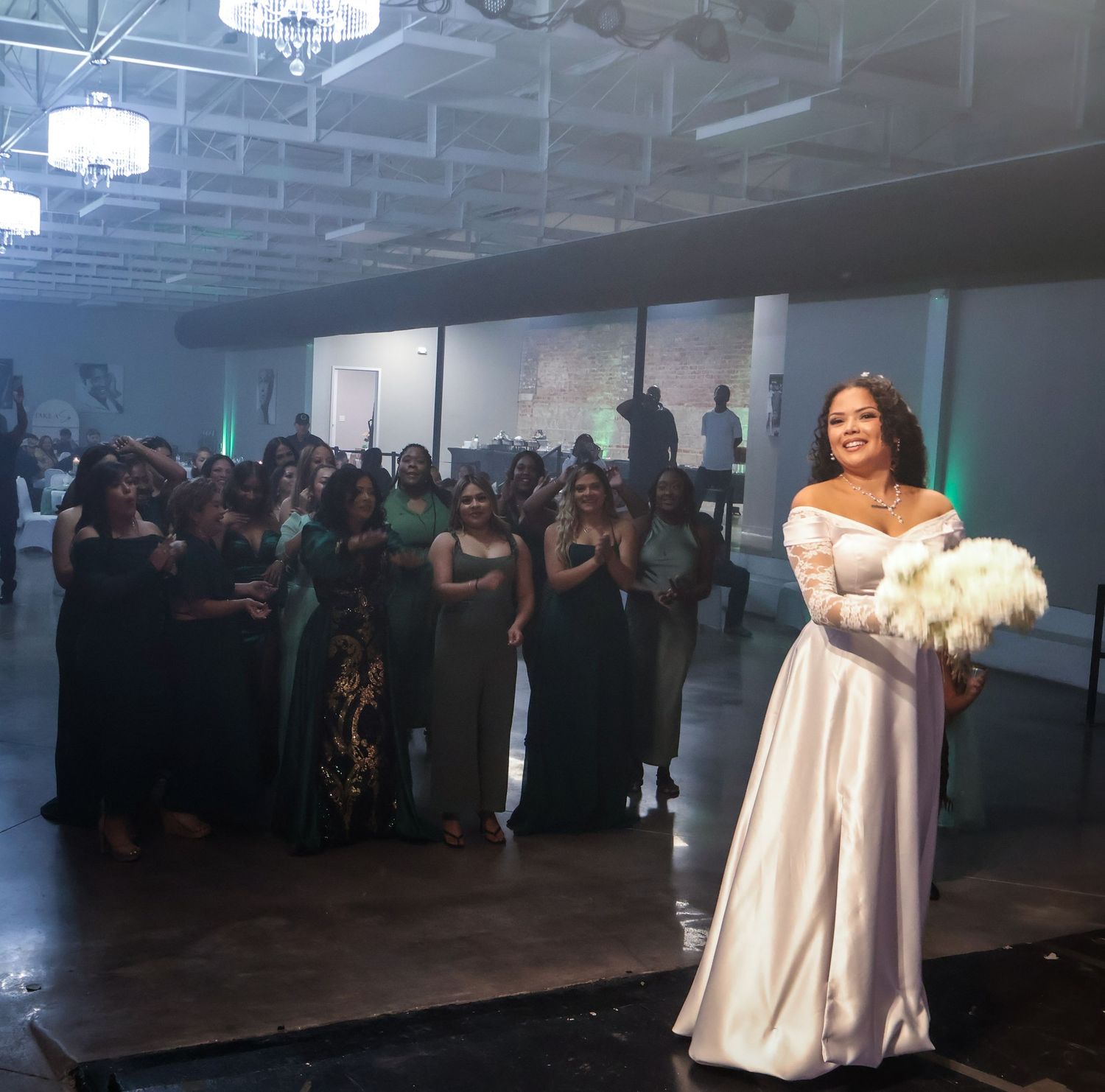 Bride in white off-shoulder gown holds bouquet before bridesmaids in green dresses at elegant wedding reception.