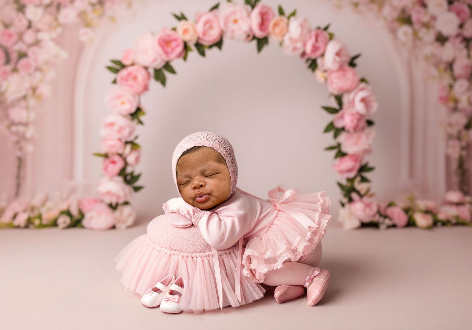 Sleeping newborn in pink dress and bonnet posed before a floral arch backdrop in studio photo.