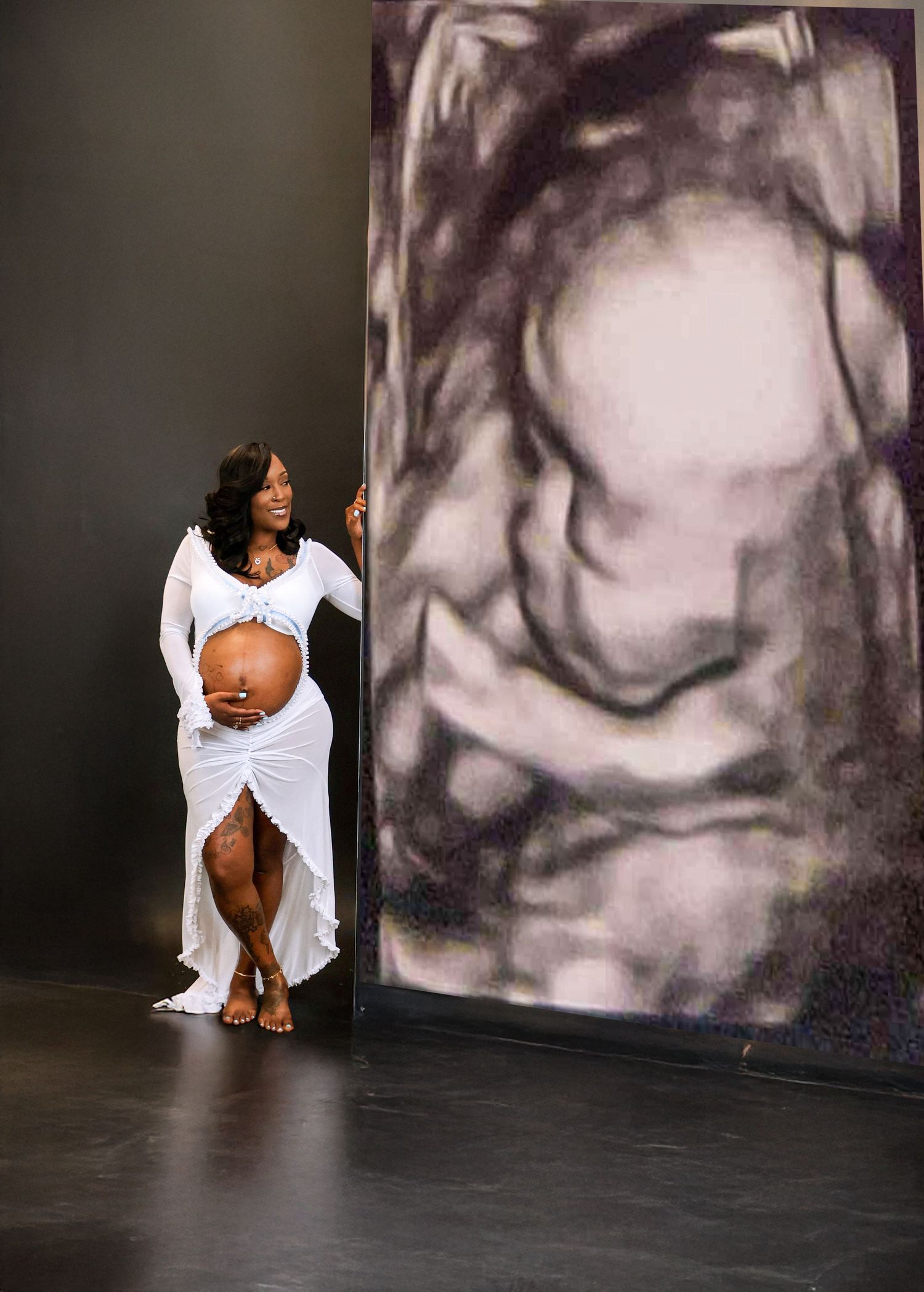 Pregnant woman in white gown poses beside large ultrasound image backdrop in professional maternity photo shoot.