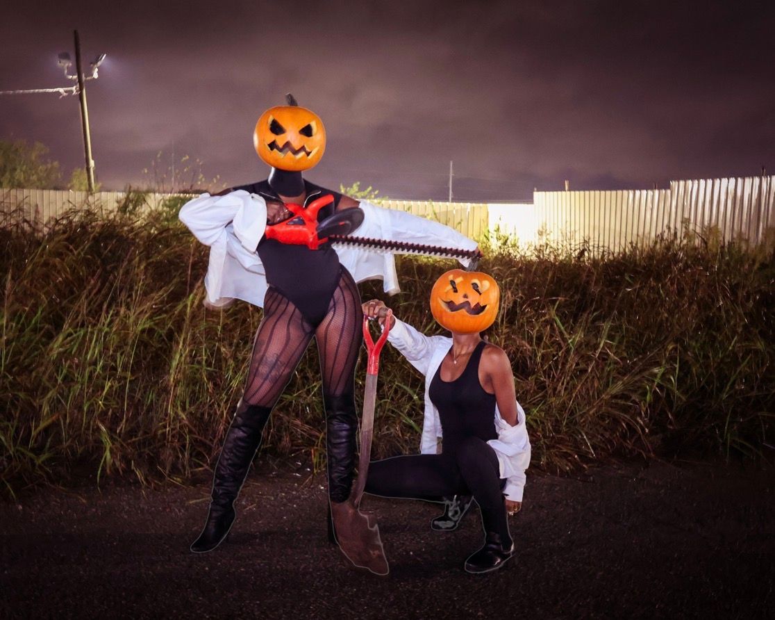 Two people in Halloween costumes wear jack-o-lantern heads while posing with a chainsaw and shovel outdoors.